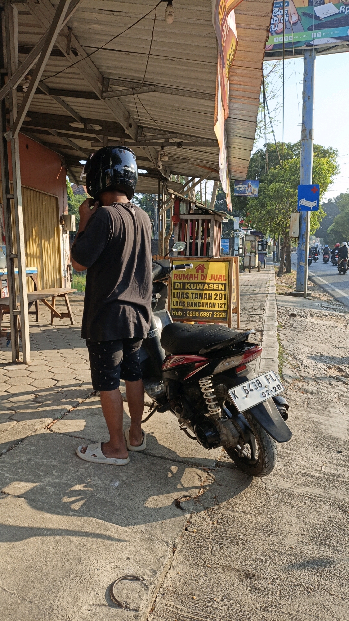 A person wearing a black helmet and black t-shirt stands next to a black and red motorcycle on a concrete sidewalk in Kuwasen, Indonesia. The person is adjusting their helmet. A yellow sign with red text advertising "RUMAH DI JUAL DI KUWASEN" (House for Sale in Kuwasen) is visible behind the motorcycle. The sign also lists land and building sizes and a contact number. A road with traffic is to the right of the sidewalk. Trees and buildings are visible in the background, and the sun casts long shadows.