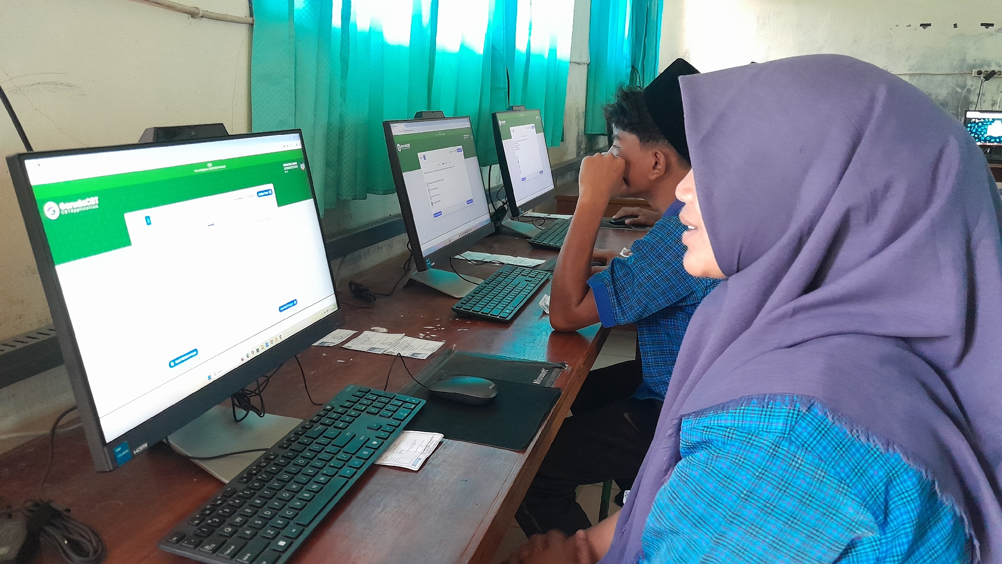 This image captures an indoor scene, likely a computer lab or classroom in Blangriek, Indonesia, during the daytime. The room features multiple computer workstations arranged on wooden desks, set against light-colored walls and bright turquoise or green curtains.
In the foreground, two individuals are visible. On the left, a young man, possibly a student, is seated at a computer, wearing a black peci (cap) and a blue plaid shirt. He appears focused, resting his right hand on his chin as he looks at the monitor. To his right, a young woman, also potentially a student, is seen in profile. She wears a purple hijab and a blue plaid shirt, similar in pattern to the man's, possibly indicating a uniform. She is looking towards the left, perhaps observing the man's screen, with a subtle, pleasant expression.
Each workstation includes a black flat-screen monitor, a keyboard, and a mouse. A black mousepad with the "Microsoft" logo is visible on the desk in the foreground, along with some papers. The monitor bezels show brand names like "HP" and "HDMI."
The main computer screen prominently displays a digital interface. A green header features a logo with the text "ComputerCBT" and "CBTapplication" below it, suggesting a computer-based test or application system. The screen shows input fields, indicating a form or login page, with visible buttons for actions like "Login" or "Submit."
The scene depicts an activity involving computer use, likely an educational assessment or an administrative application process within a focused, studious environment.