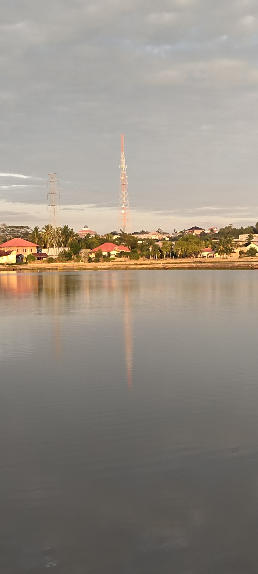 This is a photograph of the Lhokseumawe city waterfront in Indonesia. The scene depicts a wide body of calm water reflecting the sky and the structures on the opposite shore. The sky is overcast with scattered clouds. On the shore, there are several buildings with red roofs, some of which appear to be houses. Behind the buildings, palm trees and other vegetation are visible. Dominating the skyline are two tall structures: a lattice tower used for telecommunications, painted with red and white bands, and a power transmission tower with three sets of crossbars and cables. The reflection of the telecommunications tower is clearly visible in the water. The overall impression is a somewhat hazy, calm day in a coastal urban or semi-urban environment.
