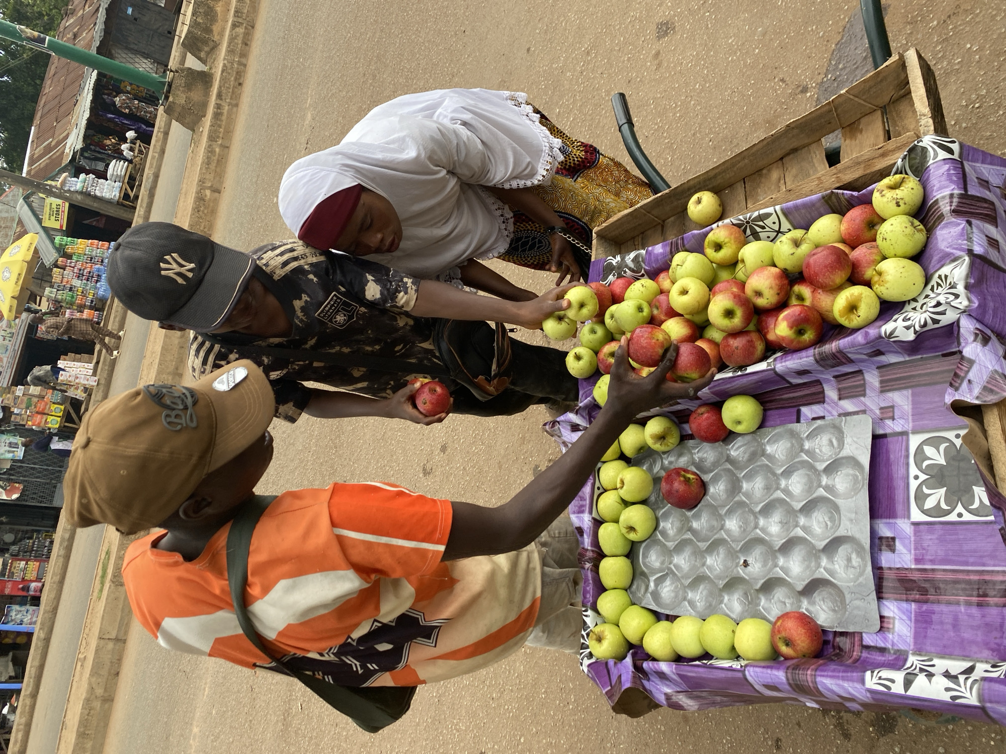 The image depicts a street scene in Bukuru, Nigeria, featuring a fruit stand with an assortment of apples. Three individuals are gathered around the stand. A young male, wearing an orange and white striped shirt and a brown baseball cap, is leaning over the cart, reaching for an apple. To his left, another young male in a black and camouflage shirt and a black baseball cap with an "NY" logo is also looking at the fruit. A woman wearing a white hijab and a colorful patterned skirt is standing on the right, also engaged with the apples. The fruit stand itself is a wooden cart covered with a purple and white striped cloth, displaying rows of green and red apples. In the background, a dusty street is visible, lined with shops and market stalls, indicative of a bustling urban environment. A yellow umbrella is also visible in the distance, suggesting a market setting.