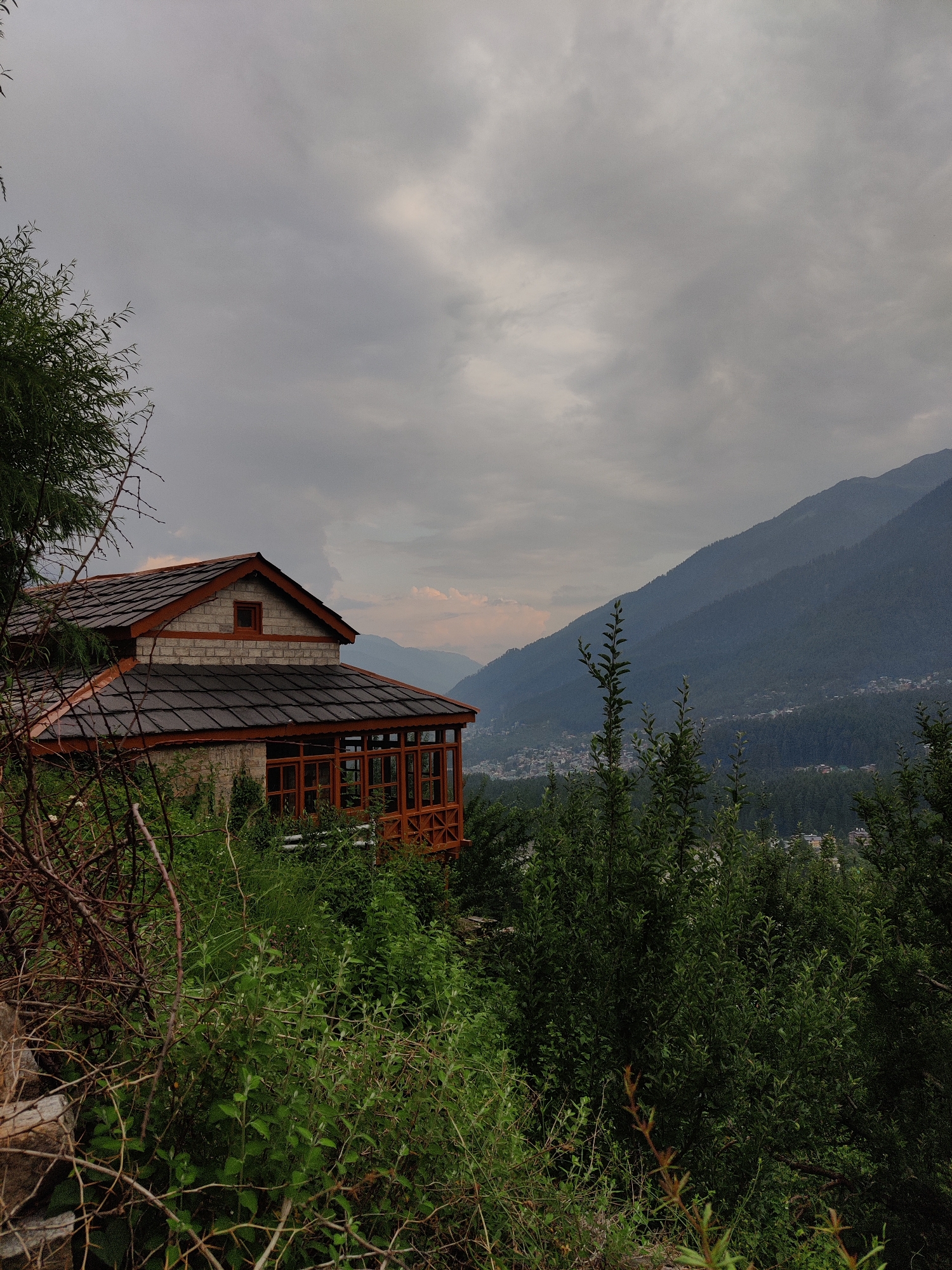 The image depicts a view of a wooden and stone house situated on a hillside in Vashisht, India. The house features a dark-tiled roof with reddish-brown trim and a covered porch with wooden beams and glass panels. The surrounding landscape is lush and green, with dense foliage in the foreground and a valley with a town nestled in the background, leading up to large, mist-covered mountains. The sky is overcast with grey clouds, suggesting a cool or rainy climate. There are no people visible in the image. The scene conveys a sense of rustic tranquility and natural beauty.