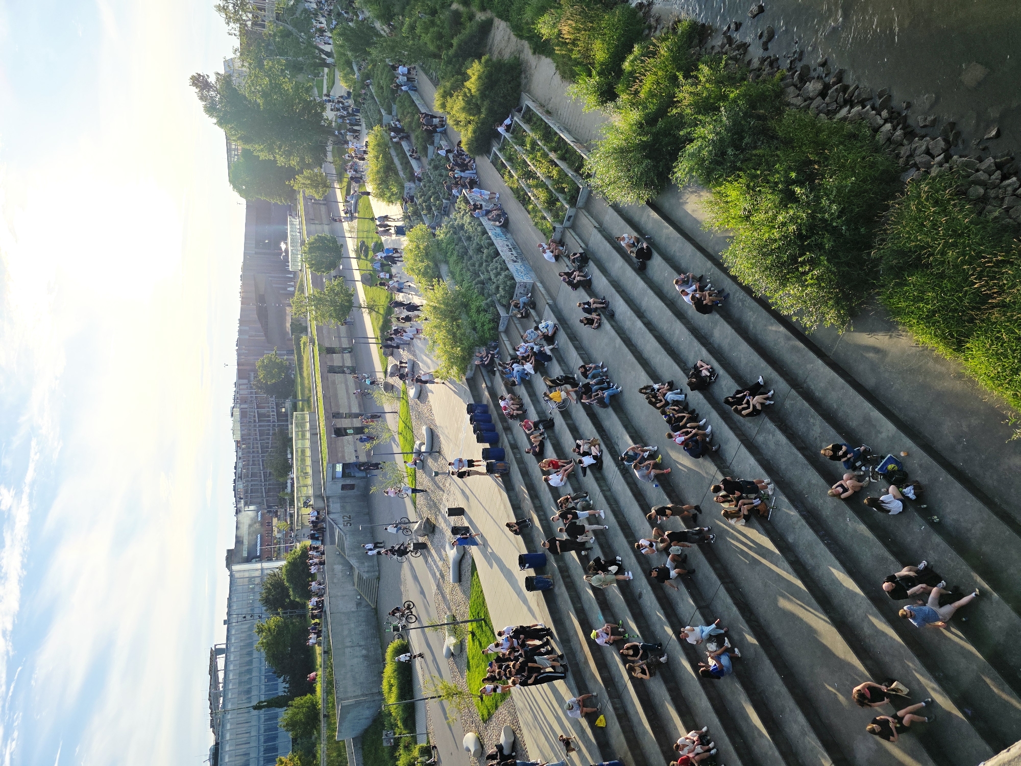 The image captures a sunny afternoon scene at the Vistula Boulevards in Warsaw, Poland. A wide set of concrete stairs descends towards the Vistula River, with numerous people scattered across the steps, seated and socializing. The steps are integrated into a landscaped area with greenery and planters.
In the background, modern glass-fronted buildings rise, indicative of the city's urban landscape. A paved walkway runs parallel to the river and the stairs, where other individuals are walking, cycling, and using scooters. The Vistula River flows gently on the right side of the image, with a rocky bank and lush vegetation lining its edge. The overall atmosphere suggests a popular recreational spot for locals and tourists alike, enjoying the warm weather and the riverside environment.