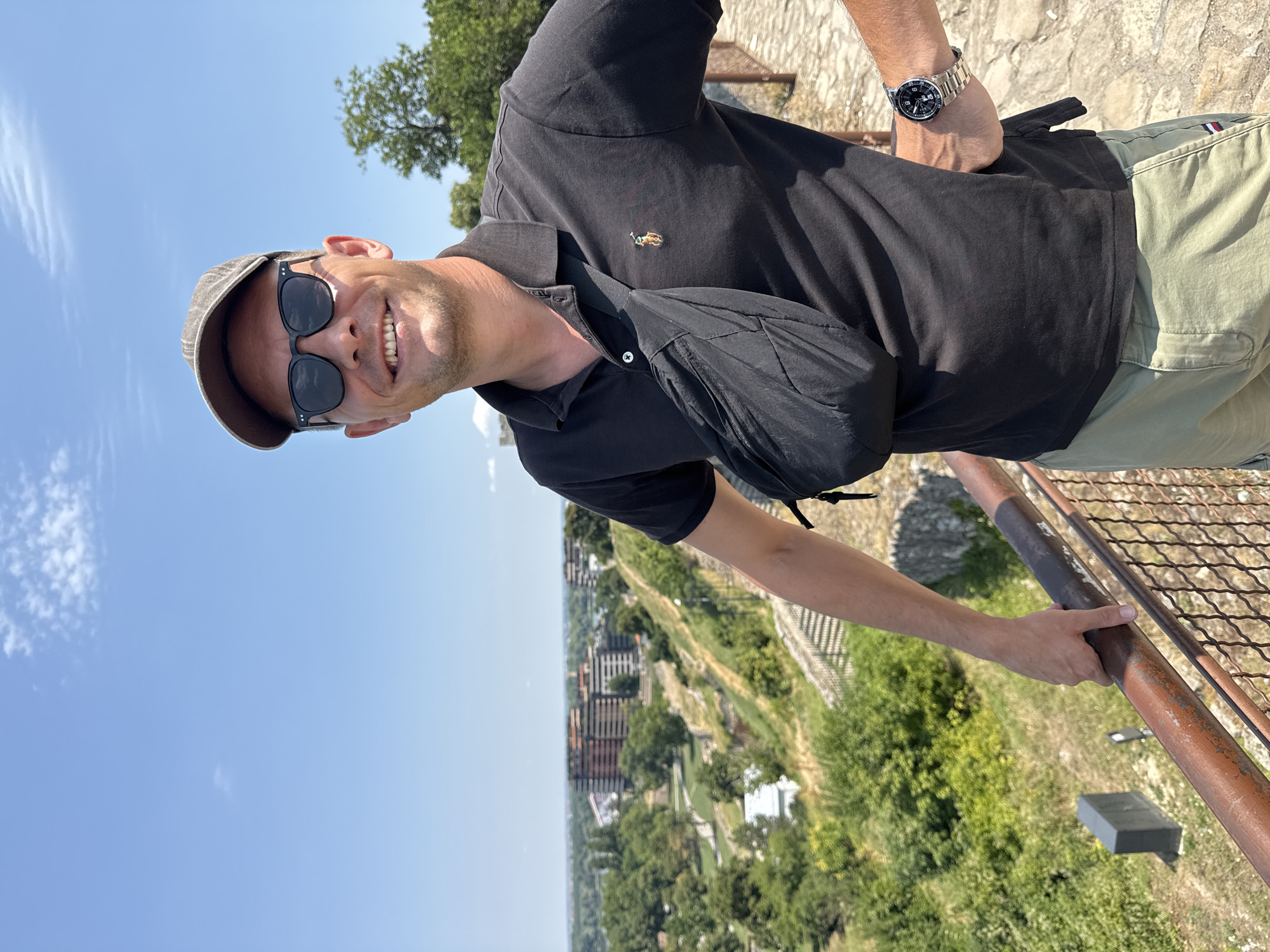 A man stands on an elevated walkway, leaning against a railing, with a city skyline in the background. He is wearing a grey baseball cap, dark sunglasses, a black polo shirt with a small embroidered logo, a black fanny pack worn across his chest, and khaki cargo shorts. A silver wristwatch is visible on his left wrist. The walkway is paved with stone and has a metal railing. In the background, the city of Belgrade, Serbia, unfolds, with green trees, buildings, and a hazy horizon. The sky is a clear blue with a few scattered clouds. The overall scene suggests a warm, sunny day in an urban park or viewpoint.