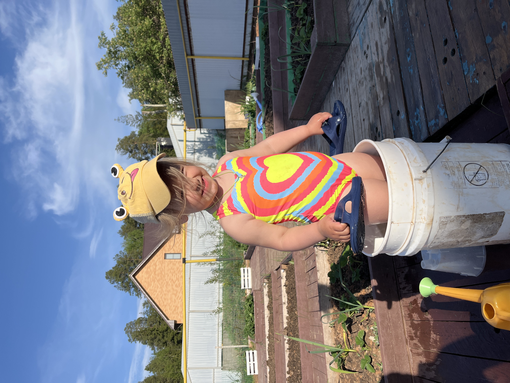 A young child, approximately 2-4 years old, stands on a wooden deck with their lower body inside a white bucket. The child wears a multi-colored swimsuit with a heart design and a yellow hat with frog-like features. They hold a pair of blue sandals in their hands.
The background shows a garden with raised wooden beds, some containing plants. Beyond the garden is a fence, trees, and a building, including a section of brick facade. A yellow watering can lies on the deck near the bucket.
The location is Chaikovsky, Russia. The weather is sunny.