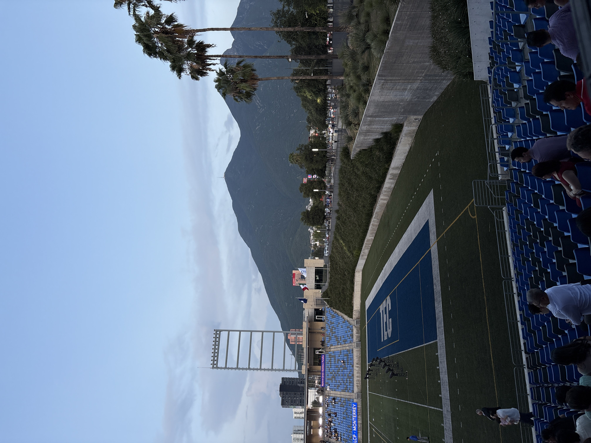 This image shows an outdoor stadium with a football field in the city of Monterrey, Mexico. In the foreground, rows of blue stadium seats are visible, some occupied by spectators. A man holding a child stands near the aisle in the left foreground. The football field is green with white markings and a blue end zone. Several spotlights are positioned on the field.
In the midground, a grandstand with blue seating is seen, with a sign that reads "Ramon Jesus Ramirez Peralta". The stadium features a tall, multi-tiered metal light tower. Behind the stadium, a street with cars and streetlights can be seen, with palm trees and other foliage lining the area. Further in the background, a large mountain range dominates the skyline under a cloudy sky. A tall, dark building and other structures are visible in the distance. Flags, including the Mexican flag, are flying in front of a building. The overall scene suggests an event is about to take place or has recently concluded.