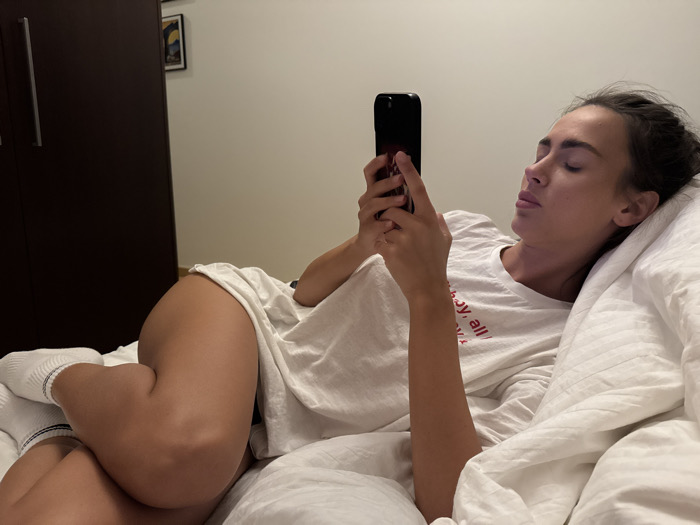 A woman is lying in a bed covered in white linens, holding a black smartphone and looking at the screen. She is wearing a white oversized t-shirt and white socks. Her legs are crossed and bent at the knees. A dark-colored wardrobe stands against the wall behind the bed. The walls are a muted light color.