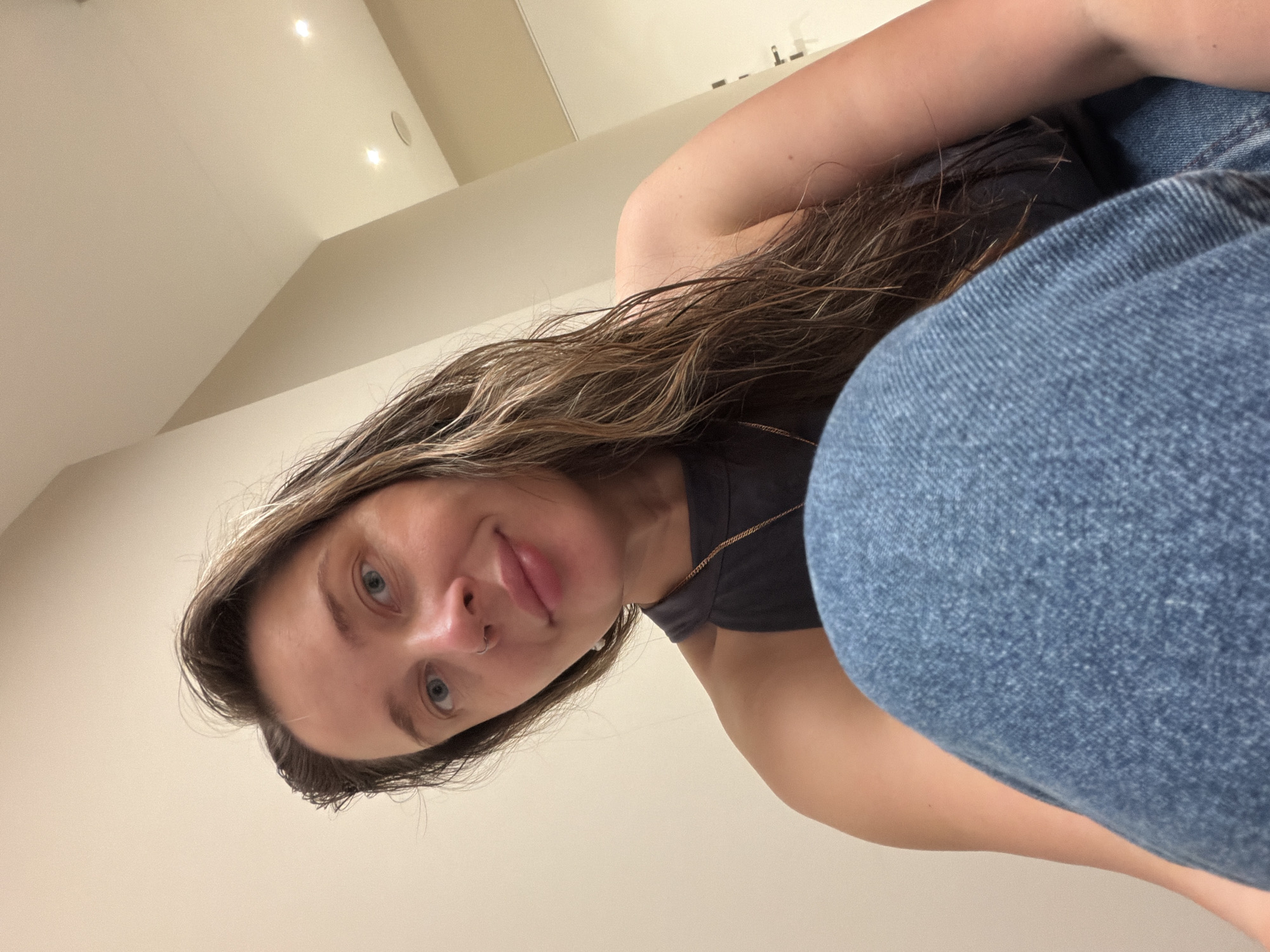The image is a close-up, low-angle selfie of a woman with long, wavy brown hair. She is wearing a dark grey tank top and blue denim jeans. Her left arm is bent, and she is looking directly at the camera with a slight smile. She has a nose ring. The background is a neutral-colored wall with recessed lighting fixtures and a doorway visible on the right. There are no visible landmarks or indicators that suggest a specific location in Moscow, Russia.