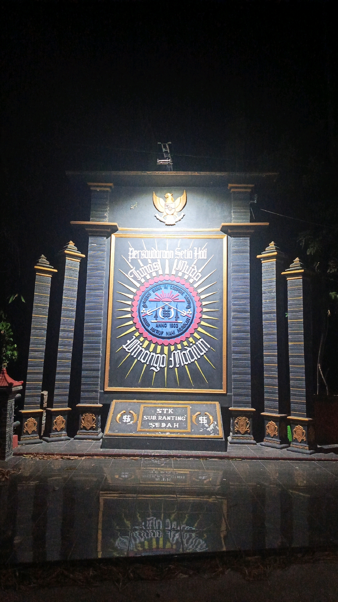 The image shows a monument at night, illuminated by artificial lights. The monument is large and ornate, with a central plaque featuring a circular emblem. The emblem contains text in Indonesian and a depiction of swords. Above the emblem is the Garuda Pancasila, the national emblem of Indonesia. Flanking the central plaque are two tall, pillar-like structures. At the base of the monument is another smaller plaque with the text "STK SUB RANTING SEDAH". The ground in front of the monument is a reflective surface, likely wet, which mirrors the monument. The surrounding environment is dark, with some foliage visible in the background, suggesting it is outdoors. The monument is located in Semanding, Indonesia.