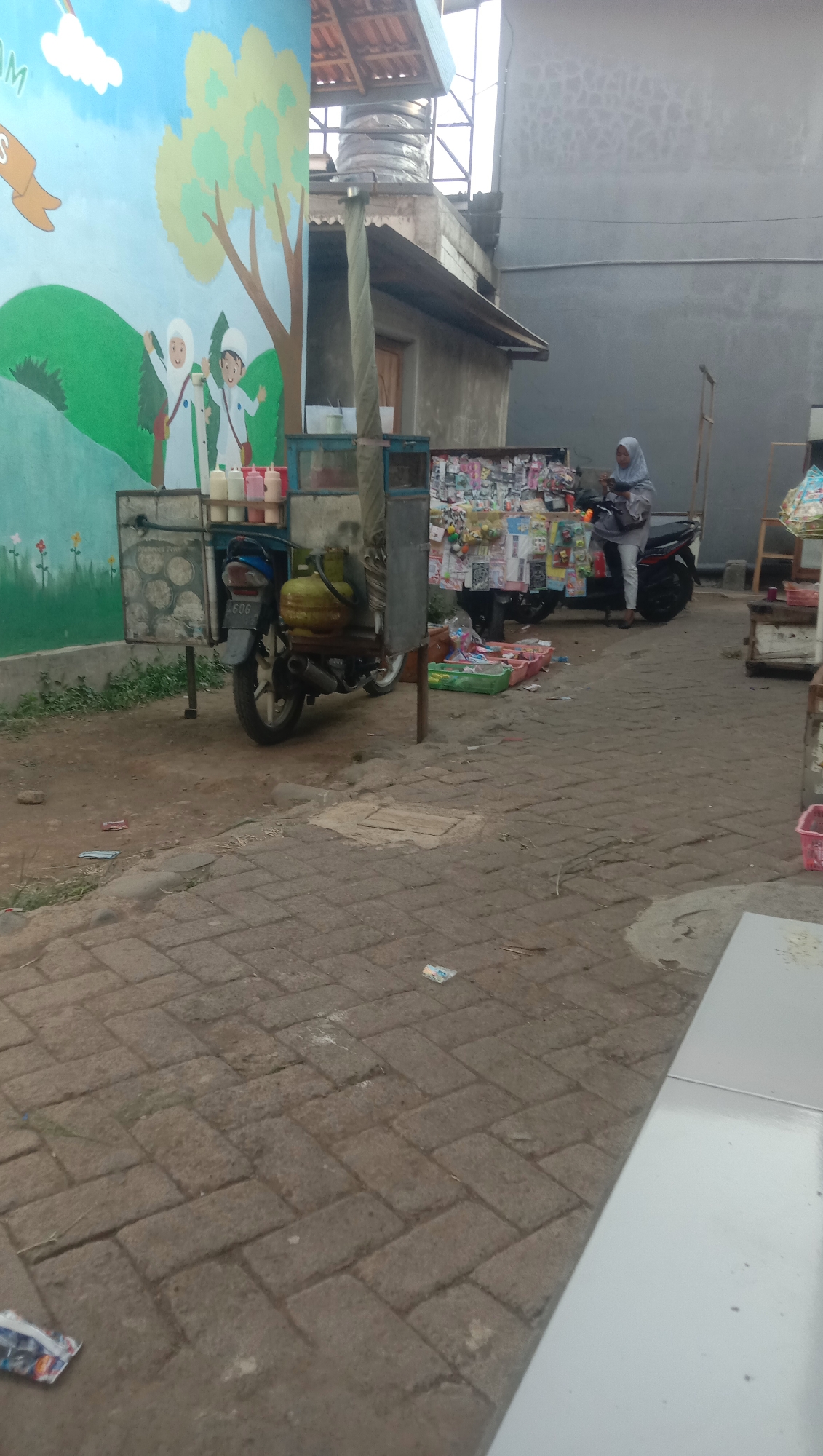 A medium shot shows a street scene with a mural on the left wall and a street vendor with a motorcycle. The mural depicts a stylized landscape with green hills, a tree, and two figures wearing hijabs. In front of the mural is a cart attached to a motorcycle, laden with bottles of colorful liquids and a gas cylinder. To the right of the cart, a woman wearing a light blue hijab and a gray outfit is standing next to a motorcycle, appearing to be interacting with the vendor's wares. The street is paved with interlocking bricks, and there are various items displayed on tables and shelves, suggesting a market or roadside stalls. In the background, buildings with gray walls and corrugated roofs are visible. The scene is likely in Karang Anyar, Indonesia, given the presence of street vendors and the architectural style.