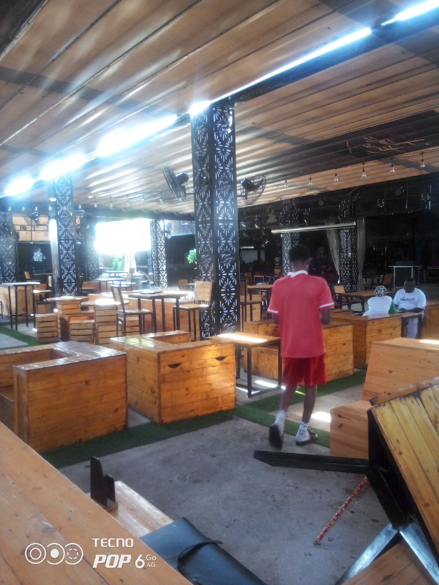 The image depicts an outdoor seating area of a commercial establishment, likely a restaurant or bar, with wooden tables and seating. The structure is covered by a slatted wooden roof with exposed lighting and beams. The environment appears to be casual, with grass-like carpeting beneath the seating areas. In the foreground, a young man wearing a red t-shirt and red shorts is walking away from the camera, towards the back of the establishment. Further back, two individuals, one wearing a white t-shirt and another in a white top, are seated at a table. The scene is illuminated by natural light filtering through the roof and artificial lighting. This image is from Howayi, Nigeria.