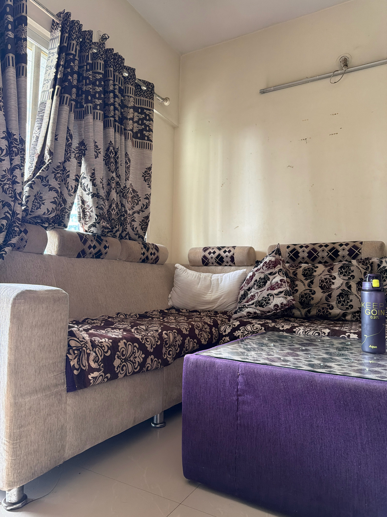 In an interior setting, likely a living room in a residential apartment in Pune, India, a beige sectional sofa with decorative patterned throw pillows and blankets occupies the foreground. A dark purple ottoman with a glass-top coffee table sits in front of the sofa. On the coffee table, a dark purple water bottle with text reading "KEEP GOING 0.97L" is visible. The room features a window with patterned curtains drawn back, revealing daylight. The walls are painted a light cream color and appear slightly aged, with some visible marks and a light fixture mounted on the ceiling. The flooring is tiled. No people are present in the scene.