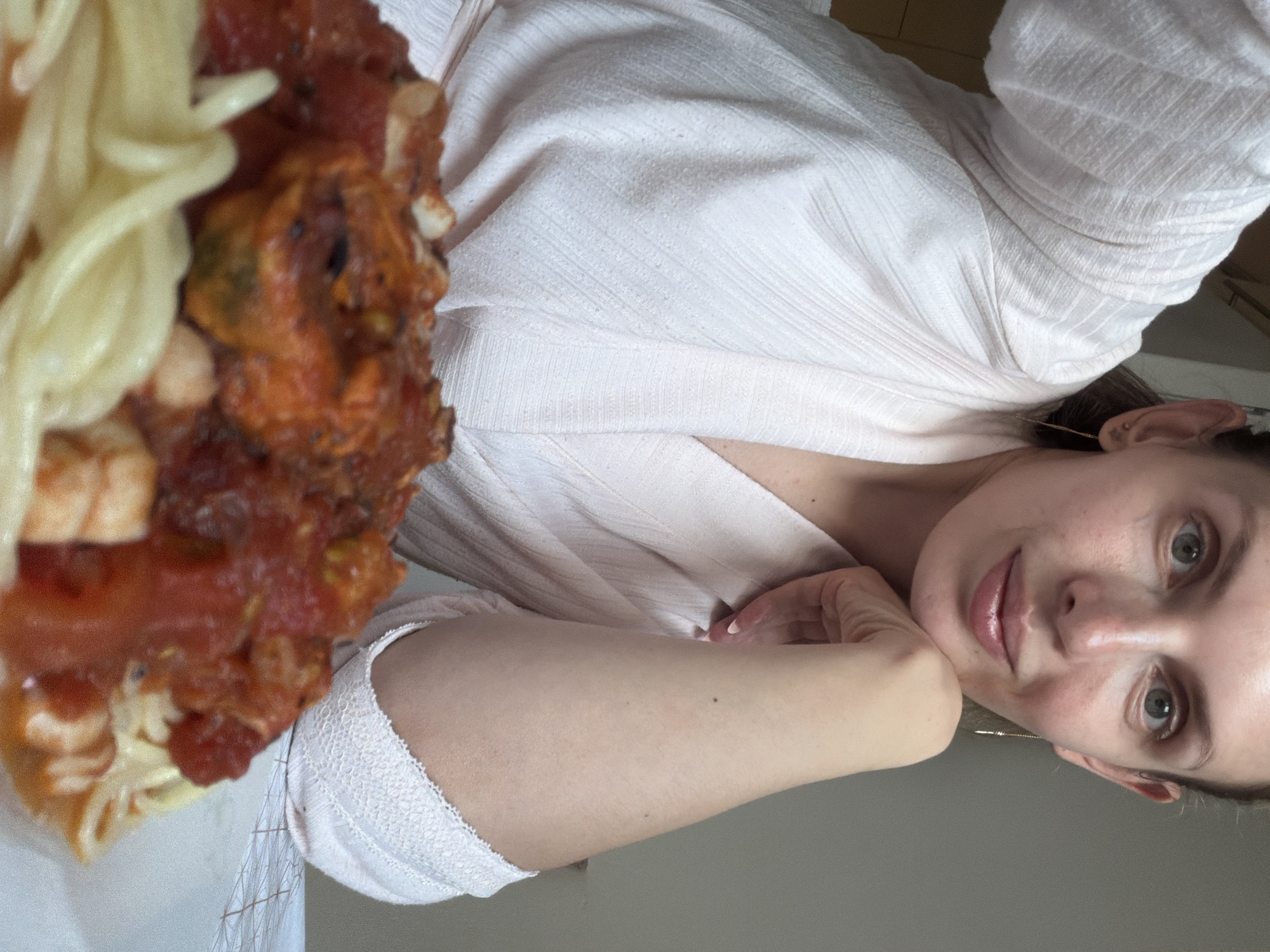 A woman is posing with a plate of pasta. The pasta is covered in a red sauce. The woman is wearing a light pink, textured shirt. The background is blurry and indistinct. There is no information in the image to indicate a location in Budva, Montenegro.