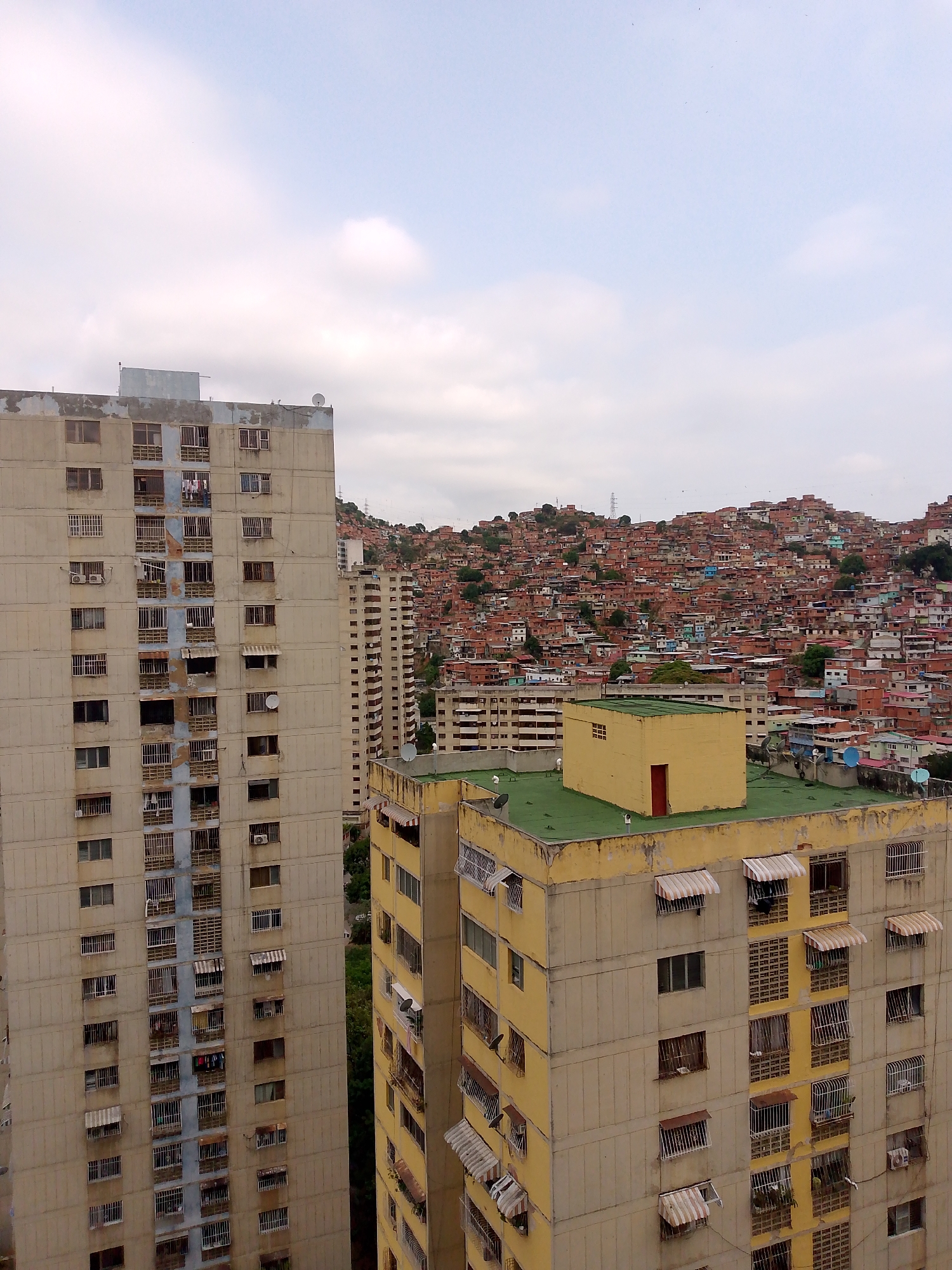 The image presents a dense urban landscape in Caracas, Venezuela, characterized by a striking juxtaposition of high-rise apartment buildings and a sprawling informal settlement on a hillside. In the foreground, two prominent multi-story apartment blocks are visible. The building on the left is a tall, concrete structure with numerous rectangular windows, many featuring security grates, awnings, and some showing clothes hanging to dry. Several satellite dishes and air conditioning units are also mounted on its facade.
To the right, another large apartment building, predominantly yellow with light-colored concrete accents, displays similar architectural details, including multiple windows with grates and striped awnings. Its flat rooftop is distinct, covered in a bright green material and featuring a small, box-like yellow structure with a red door.
Beyond these structured apartment buildings, a vast hill rises, densely covered from its base to its summit with countless small, multi-colored houses and shacks, typical of a "barrio" or favela. These informal dwellings appear tightly packed, creating a mosaic of roofs and walls that defines the mountainous backdrop. Utility poles or towers are faintly visible on the distant ridge.
The sky above is overcast with diffused clouds, indicating a daytime scene, likely late morning or afternoon. No direct human activity is visible, but the architectural details suggest ongoing daily life within the residential structures. The scene powerfully conveys urban density and the layered socioeconomic fabric characteristic of Caracas. No discernible text is visible in the media file.