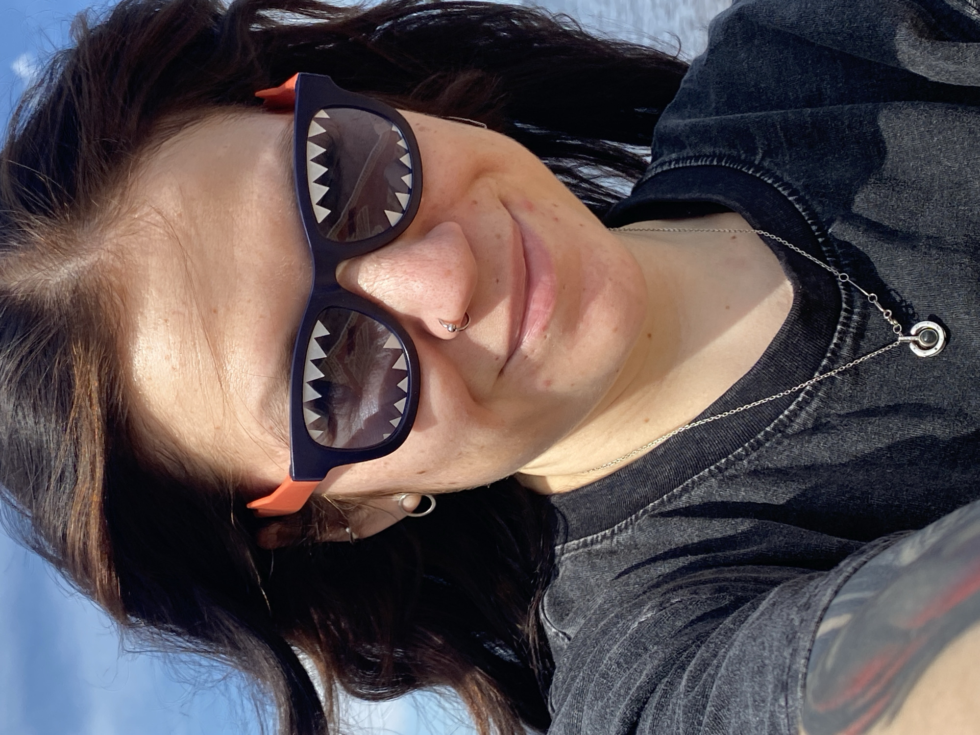 A woman is facing the camera, wearing sunglasses with a jagged pattern on the lenses, a nose ring, and a silver necklace. She has dark hair and is wearing a dark t-shirt. The background shows a hint of blue sky and what appears to be water. It is not possible to verify the location as Chaikovsky, Russia based on the provided image.
