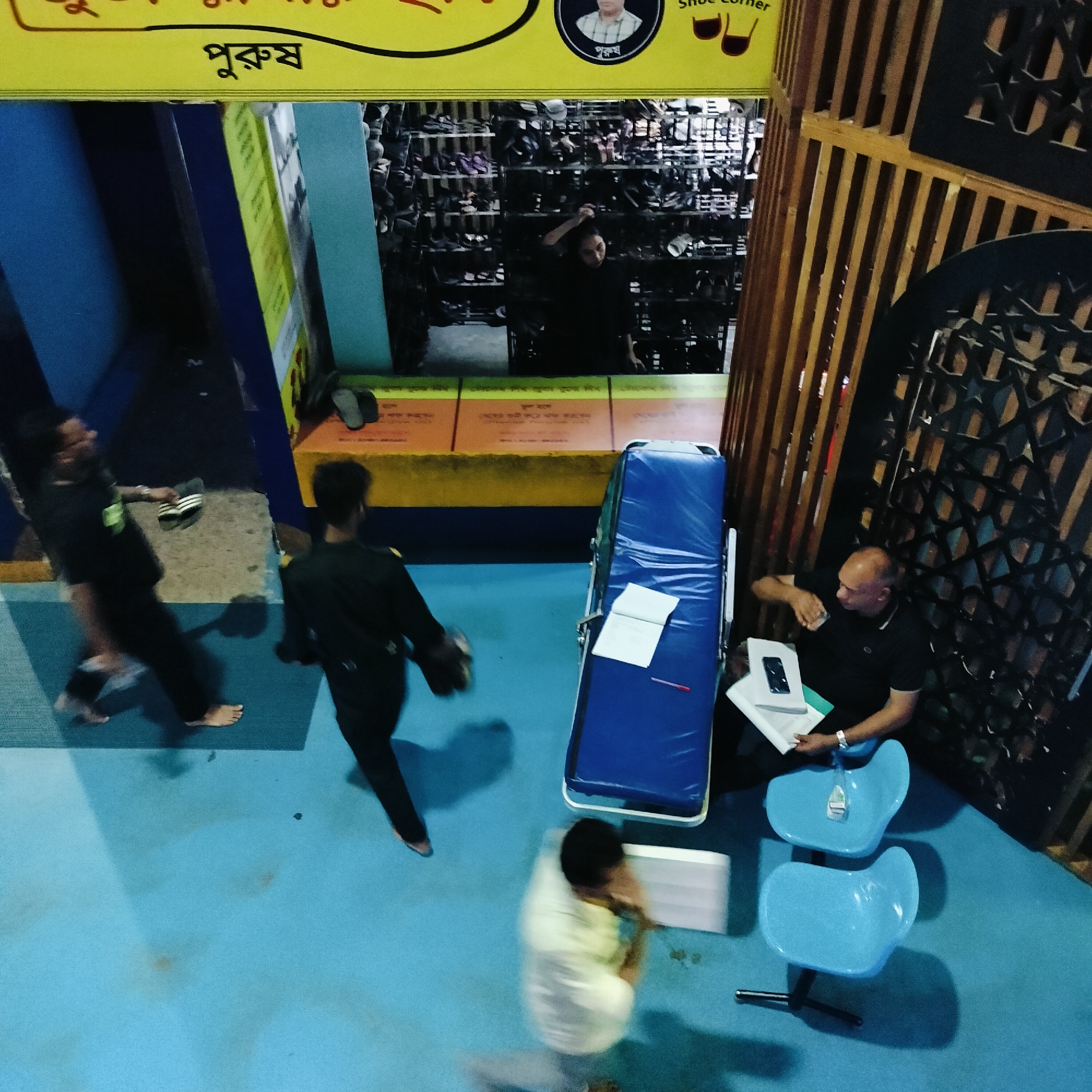 A high-angle shot shows the entrance to a shoe store, a blue-floored waiting area, and several people at an establishment in Dhaka, Bangladesh.
The shoe store has a yellow sign above the entrance; visible lettering in Bangla. Inside, shelves filled with shoes can be seen, and a woman is reaching up to adjust her hair.
In the waiting area, a man sits on a blue chair, holding papers and a phone. Two empty blue chairs are nearby. A blue padded examination or waiting bed stands between the man and the shoe store entrance.
Three people are walking across the blue floor in the direction of the camera. One is barefoot, carrying sandals. The second is carrying shoes. The third is blurred, suggesting rapid movement.
The environment consists of a combination of a shoe store with wooden trim to the right, and a yellow and blue corridor leading into the building on the left.