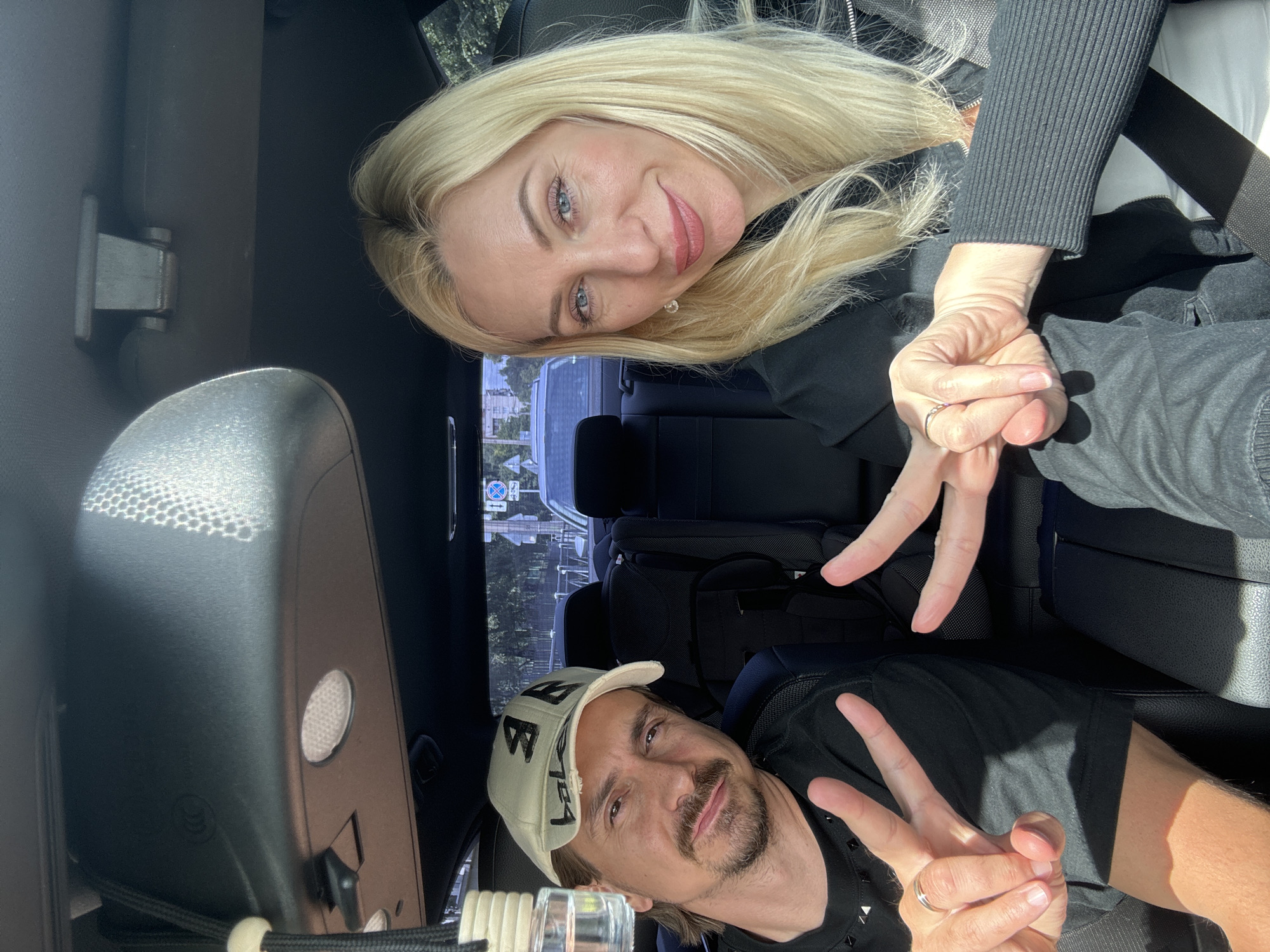 Two individuals, a man and a woman, are seated inside a car, appearing to be in the passenger and driver's seats respectively, based on their positioning. The man, wearing a black t-shirt and a beige baseball cap, is looking towards the camera and making a peace sign with his right hand, while his left hand is also raised with two fingers extended, also forming a peace sign. He has a mustache and goatee. The woman, with blonde hair and blue eyes, is wearing a black top with a white zipper visible. She is also making a peace sign with her right hand, which is positioned closer to the camera. Her left hand is clasped with the man's right hand. The car's interior is visible, including the rear seats, a car seat, and the rearview mirror, which reflects sunlight. Outside the car, trees and buildings can be seen, suggesting an urban or suburban environment. A car window on the right side of the frame shows a street sign, but it is not legible enough to identify the location. The lighting suggests it is daytime.