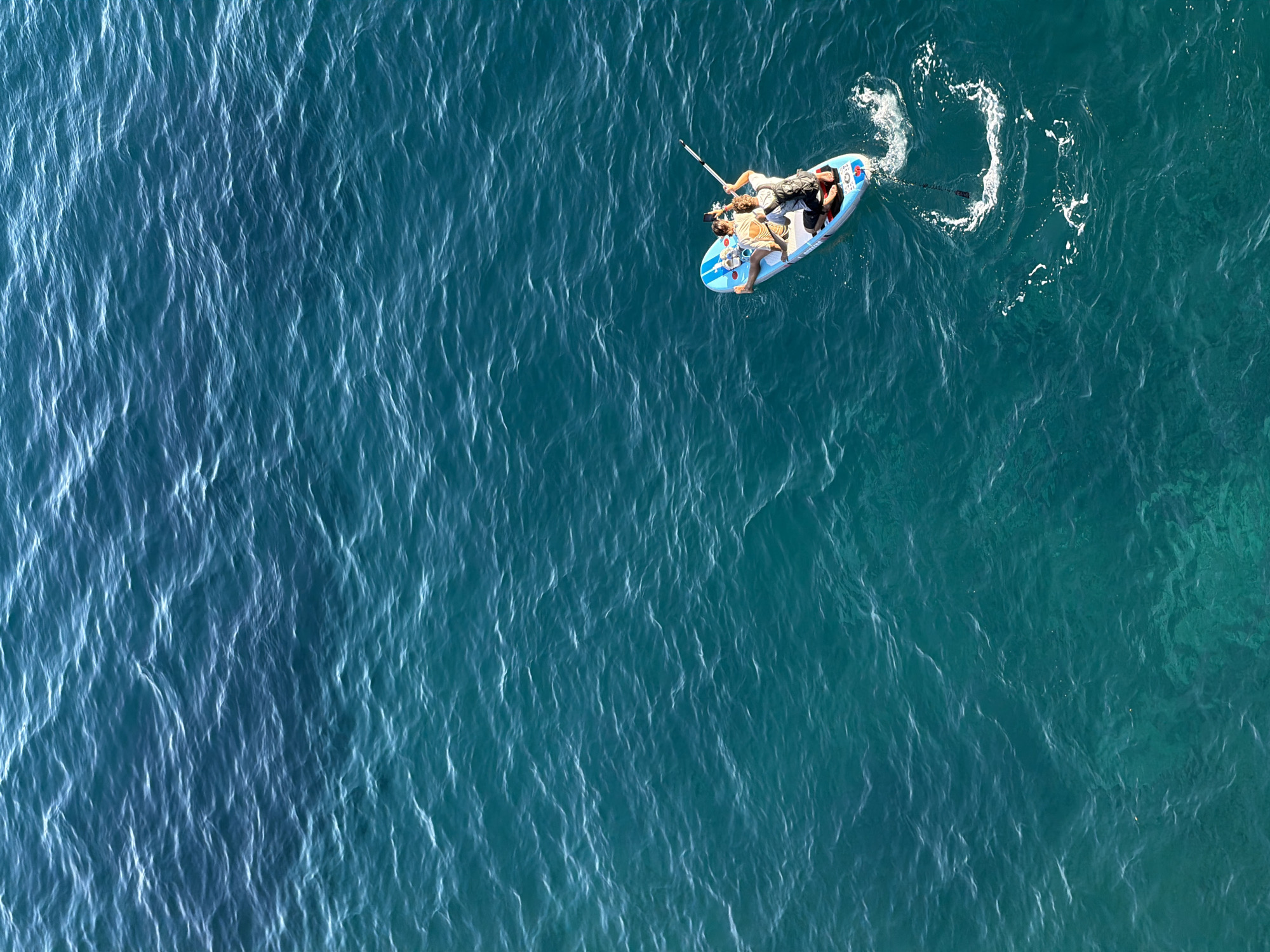 An aerial, high-angle, long shot shows two people on a paddleboard in the turquoise water of Petrovac na Moru, Montenegro. The paddleboard is decorated with a blue and white design and has red accents. The person in the back is paddling with a black and white paddle. The person in the front is sitting and looking down at a phone. Both individuals are wearing light-colored clothing. The water is calm with small ripples, and the sun is casting highlights on the surface. There is a wake behind the paddleboard, indicating movement through the water. The vast expanse of the sea fills the majority of the frame, with a gradient of blues from a darker shade at the top to a lighter, more vibrant turquoise at the bottom.