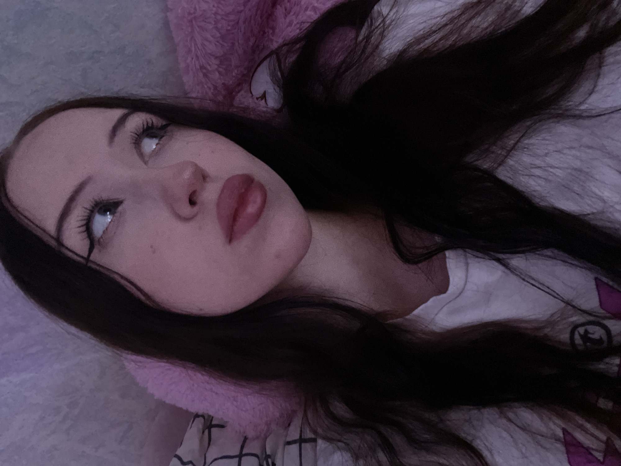 A young woman with long, dark hair, wearing a white t-shirt, is looking to the right. She has dark eyeliner and mascara on, and her lips are painted with a pinkish-red lipstick. She is lying down, possibly in a bed, with a pink fluffy blanket or pillow behind her. The background appears to be a wall with a subtle texture, illuminated by soft, purplish light. The overall impression is of a casual, intimate setting. There is no information in the image that would suggest a specific location within Votkinsk, Russia.