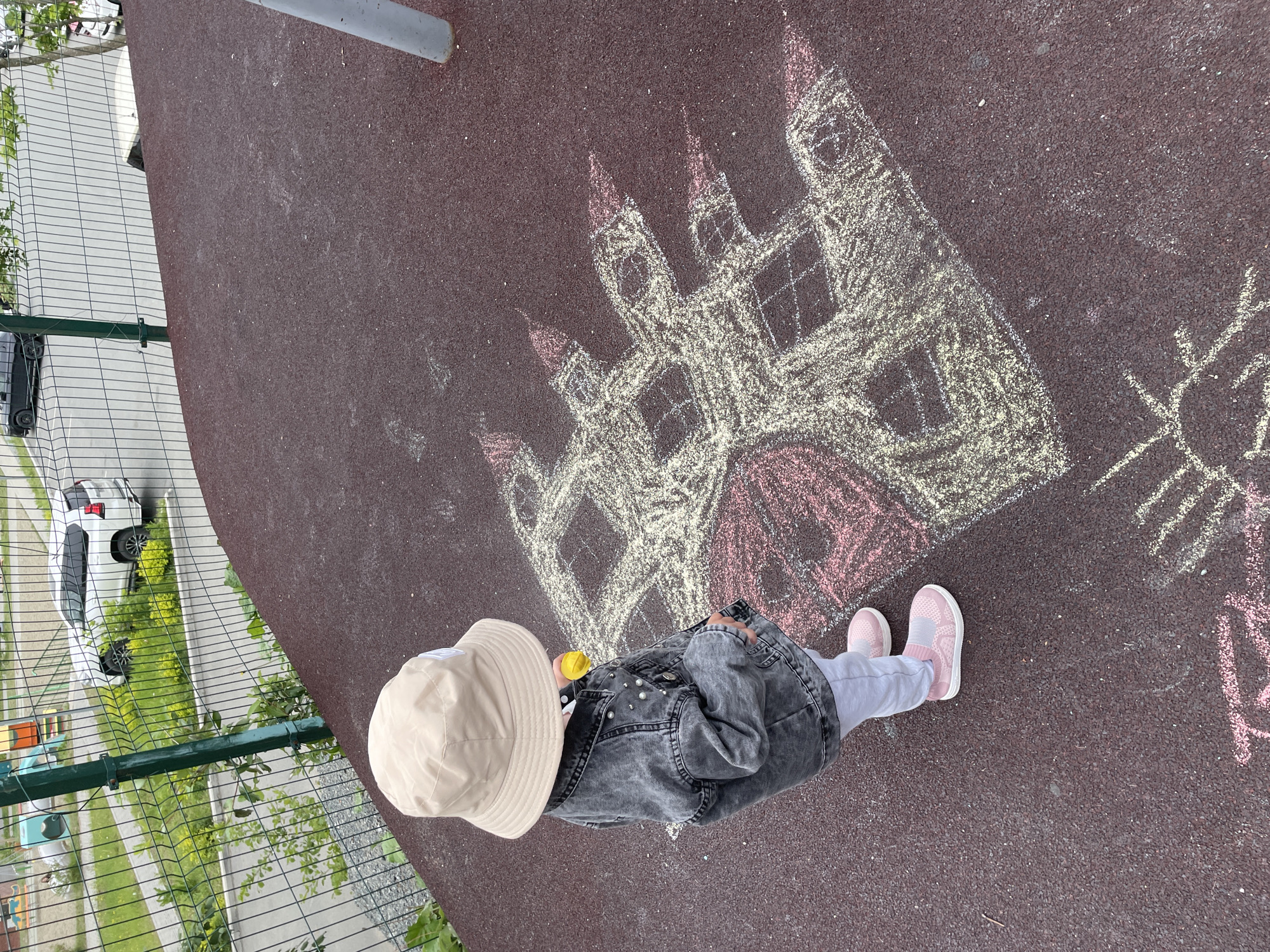 A child wearing a tan bucket hat, a grey denim jacket, light grey pants, and pink sneakers is drawing with chalk on a red surface. The chalk drawing depicts a castle with red accents. The scene appears to be an outdoor area, possibly a playground, with a metal fence visible on the left side of the image. Beyond the fence, there is greenery and parked vehicles. The photo was taken from a high angle, looking down at the child and the drawing. The image does not contain specific data suggesting that the image was taken in Vladivostok, Russia.