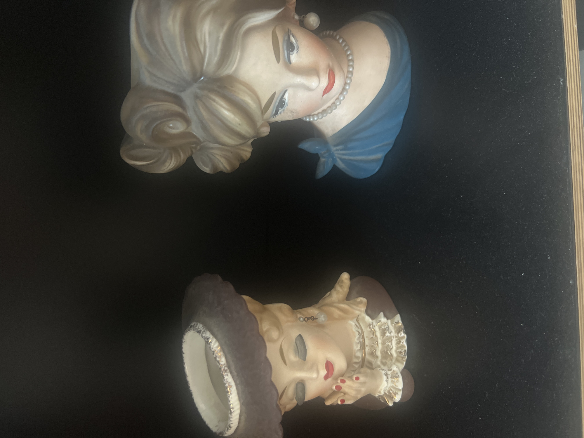 The image shows two ceramic figurines of women's busts displayed on a black surface, likely a shelf, against a black background. Both figurines are depicted in a vintage style. The figurine on the left wears a brown hat with a decorative rim and a ruffled collar, with a hand near its chin. The figurine on the right is adorned with a blue off-the-shoulder garment and a pearl necklace. The scene appears to be an indoor setting. The image was taken on April 16, 2024, at approximately 12:49 PM. The location where the photo was taken is Hadera, Israel.