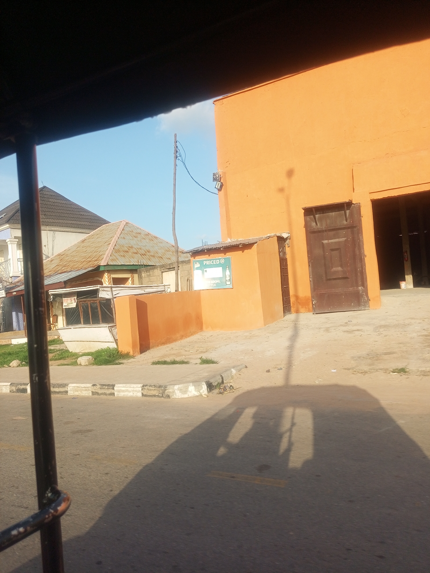 The image captures a street scene in Jalingo, Nigeria, likely in the late afternoon or early evening, given the long shadows cast by the structures and an unseen vehicle. The dominant colors are the warm orange of a building and the blue of the sky.
The foreground is occupied by the shadow of a vehicle, possibly a tricycle or a rickshaw, suggesting the perspective is from inside such a vehicle. A dark metal bar from the vehicle's structure is visible on the left.
In the mid-ground, there's a street with a concrete curb and asphalt. Beyond the curb, a small structure with a white awning and glass windows is visible, with an orange-colored wall extending from it. A sign with text and a picture of a bottle is attached to this orange wall. Further back, a building with a rusty, corrugated metal roof stands out. To the right, a large, imposing orange building with a dark brown, wooden door dominates the scene. The entrance to this building appears to be an open bay. A utility pole with wires is also present, contributing to the urban setting.
The scene appears to be in a town or city, with a mix of residential and commercial structures. The weather seems clear with a bright sky. There are no people visible in the frame. The overall impression is one of a typical day in a Nigerian urban environment.
Visible text includes "PRICED @" and "500ML BOTTLE" on the sign attached to the orange wall.