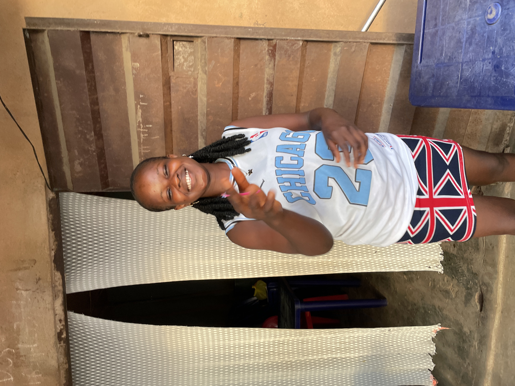 A young Black woman is standing in front of a wooden door and white curtains. She is wearing a white basketball jersey with the word "CHICAGO" and the number "23" in light blue. She also has a skirt with a Union Jack pattern. Her hair is braided and she is smiling and pointing at the camera. To the left of the woman, through the white curtains, two blue chairs and a red chair are visible. A blue plastic table is on the right side of the image. The wall above the woman has some chalk marks. The scene appears to be outdoors, possibly in a market or shop area in Nkpor, Nigeria.