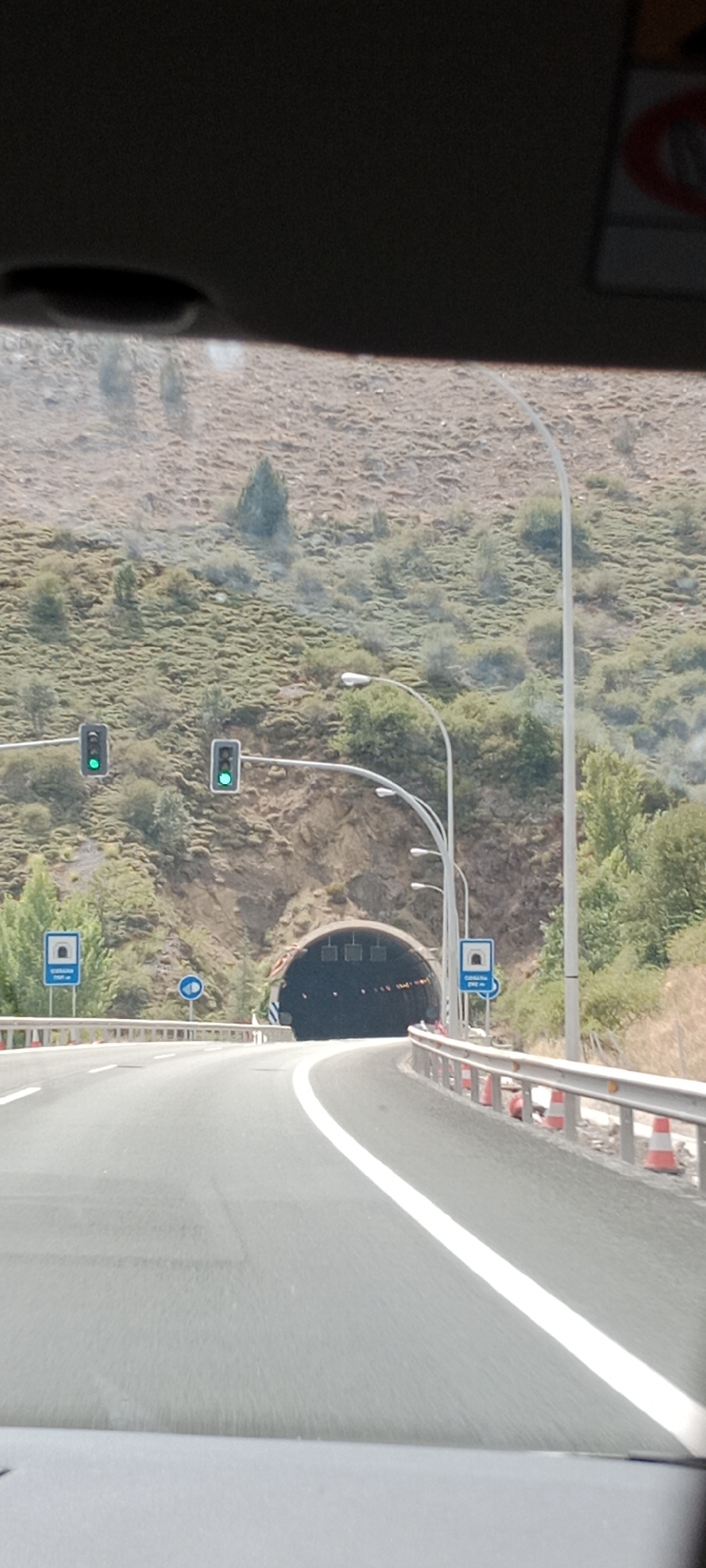 The image shows a view from inside a vehicle approaching a tunnel entrance. The road ahead is paved and has a white line marking the lane. On the left side of the road, there are two traffic lights displaying green, and a blue sign with a white tunnel icon and text below it. On the right side of the road, there is a metal guardrail with orange traffic cones placed behind it, and a light pole with multiple lights. Beyond the road and guardrail, there is a rocky hillside covered with sparse vegetation. The tunnel entrance is a dark, arched opening in the rock face, illuminated by internal lights. The sky is not visible. The location is identified as San Esteban del Molar, Spain.
