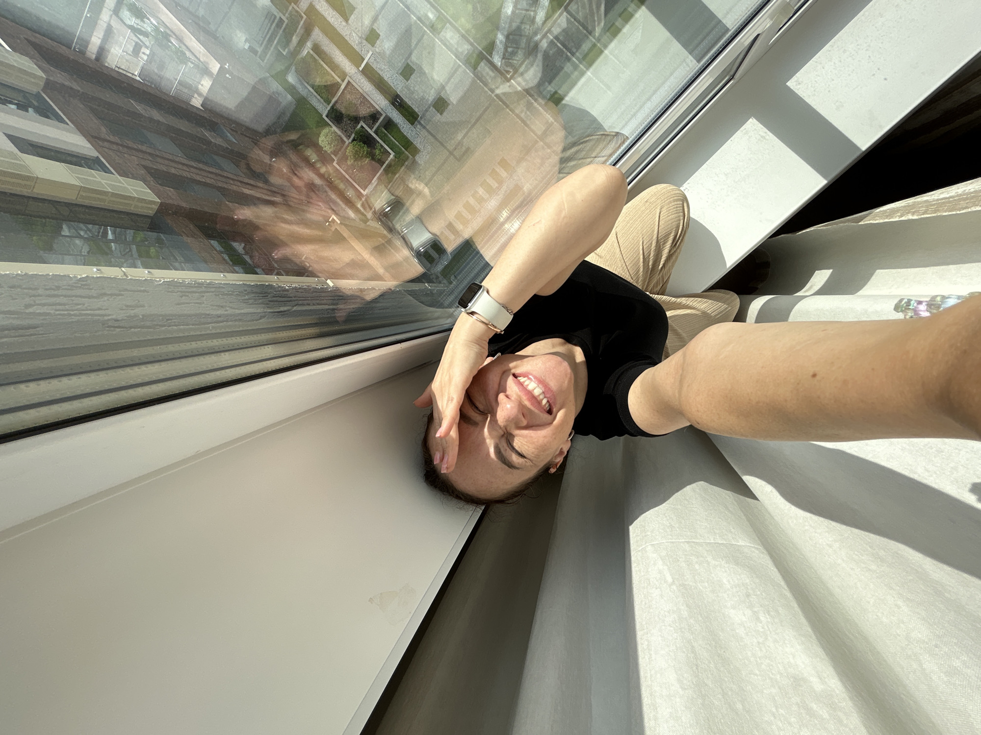A woman with her hand on her forehead smiles brightly, her eyes closed in a gesture of joy or exasperation. She is wearing a black t-shirt and beige pants, and a white smartwatch on her left wrist. The woman is positioned indoors, with a large window to her right reflecting a building and courtyard outside, suggesting an urban environment. The lighting indicates it is daytime, with sunlight casting shadows. The overall scene is a selfie taken from a high angle, looking down at the woman. Based on the architectural style of the reflected building, it is plausible this image was taken in Yekaterinburg, Russia, a city known for its diverse architectural landscape.
