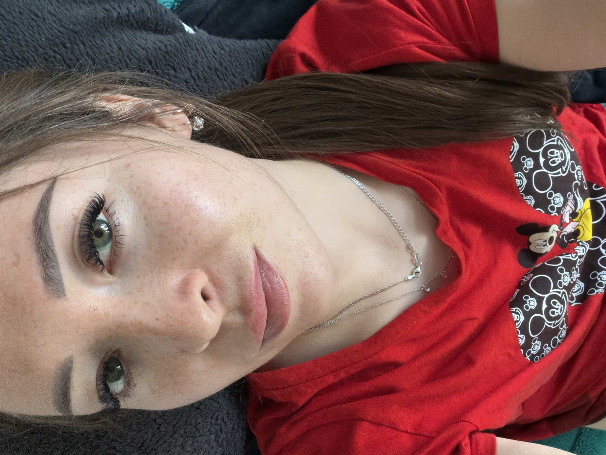A close-up, overhead selfie shows a young woman with green eyes and freckled skin. She has dark, arched eyebrows and long, dark eyelashes. Her lips are full and appear to be lightly colored. She is wearing a red t-shirt with a Mickey Mouse graphic on it and a silver necklace. A small, diamond-like earring is visible in her right ear. Her brown hair is pulled back into a ponytail. The background appears to be a dark, textured blanket. The image was taken indoors, and based on the context of the provided location, it is likely in Tujmazy, Russia.