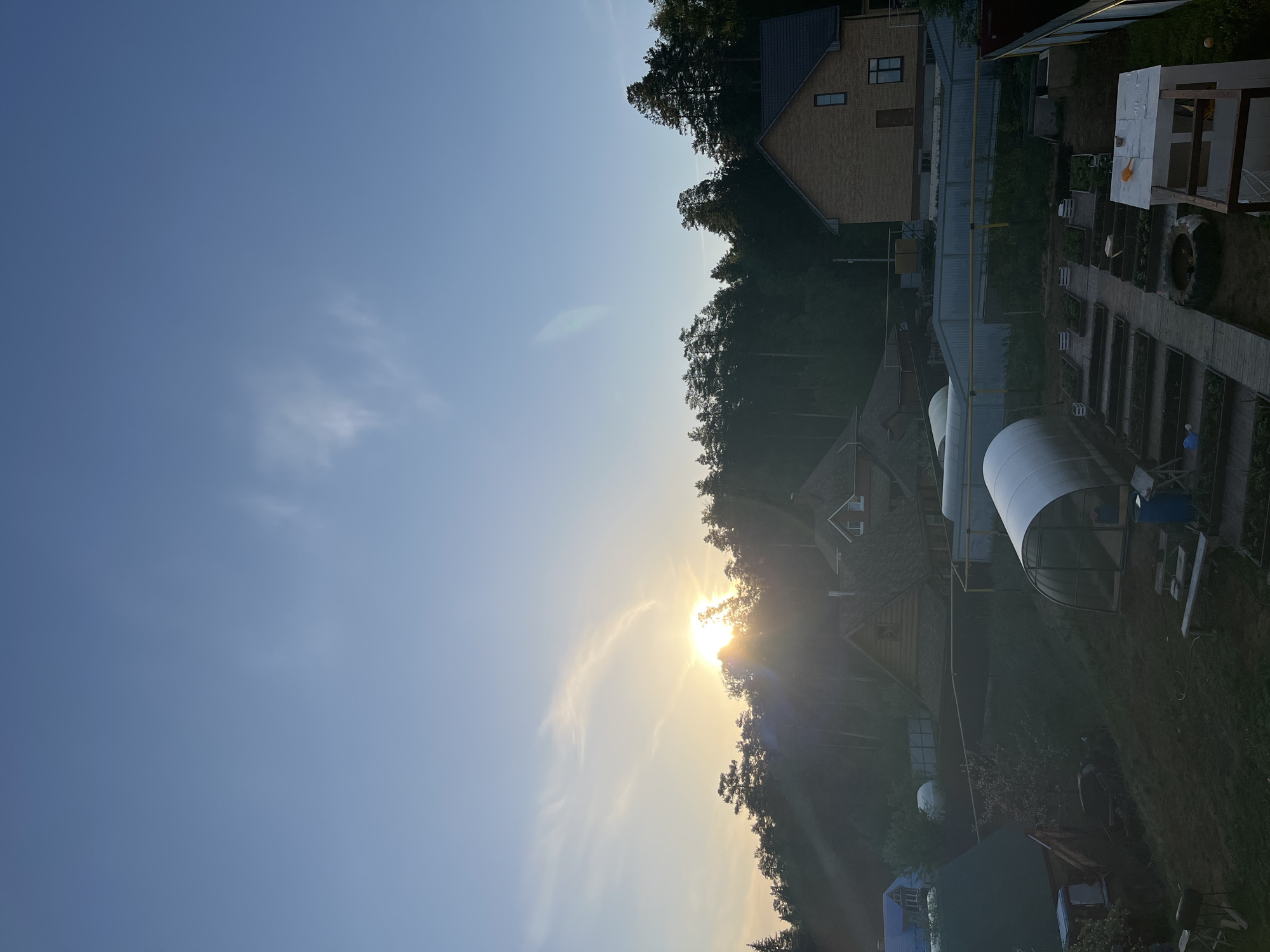 The image shows a landscape with the sun low in the sky, partially obscured by trees and clouds. The sky is a gradient of blue to light yellow near the horizon. Below the horizon line, there is a dense line of trees with buildings interspersed, including a brick house with a dark roof, a greenhouse with a white arch, and other structures. A garden with raised beds and a wooden walkway is visible. Based on the location provided, the image depicts a scene in a residential or rural area of Chaikovsky, Russia, possibly taken at sunrise or sunset. No people are visible. The focus is on the natural landscape and built environment within a residential area.