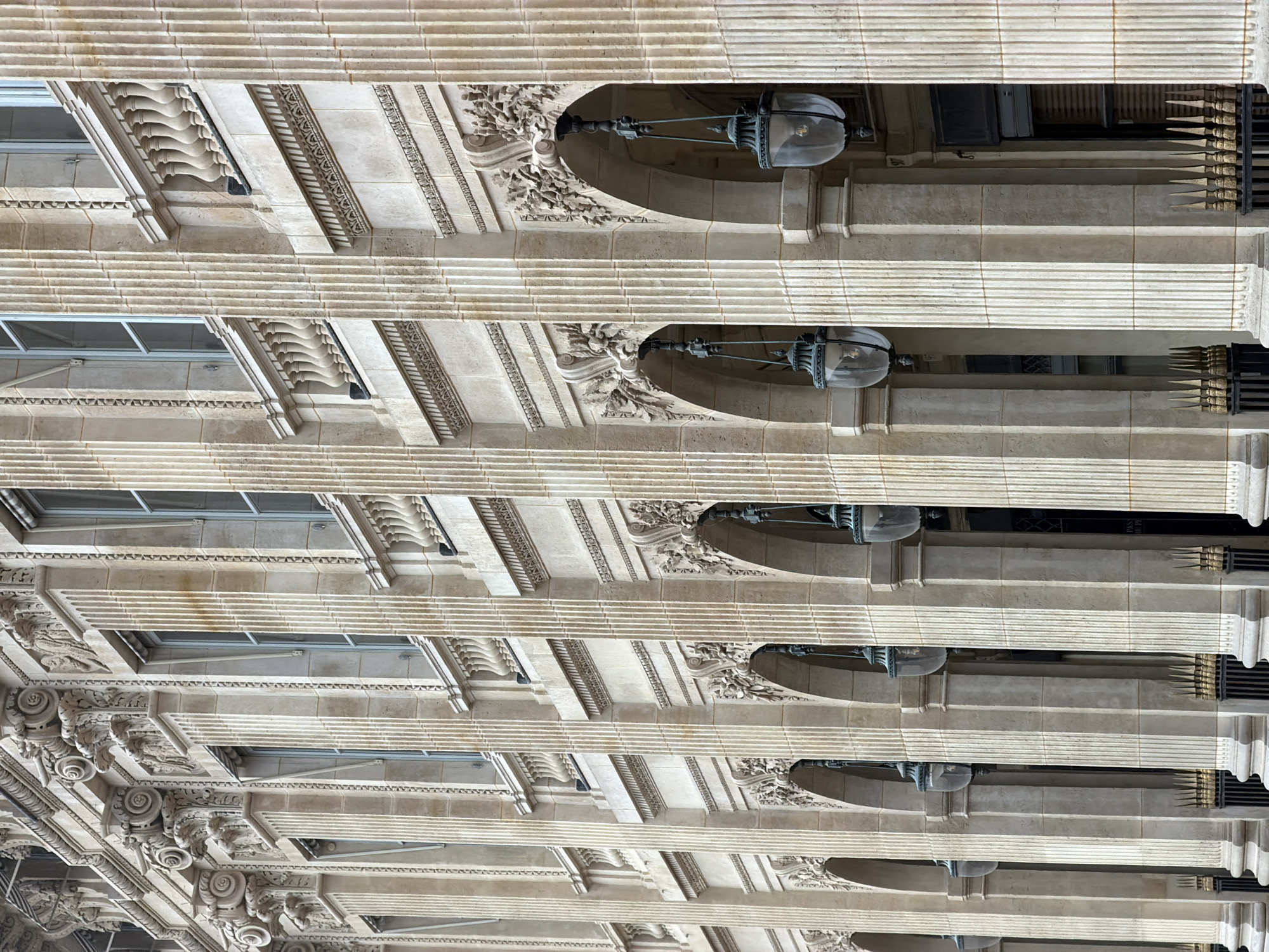 The image is a perspective shot looking up at the exterior of a classical-style building. The location appears to be in Paris, France, given the architectural style. The building features ornate details such as fluted columns, decorative carvings, and corbels. There are visible windows and antique-style lanterns hanging from the facade. The building's material seems to be a light-colored stone. The lighting is even, suggesting daylight, and there is no visible presence of people. The viewpoint emphasizes the repetitive pattern of the architectural elements.