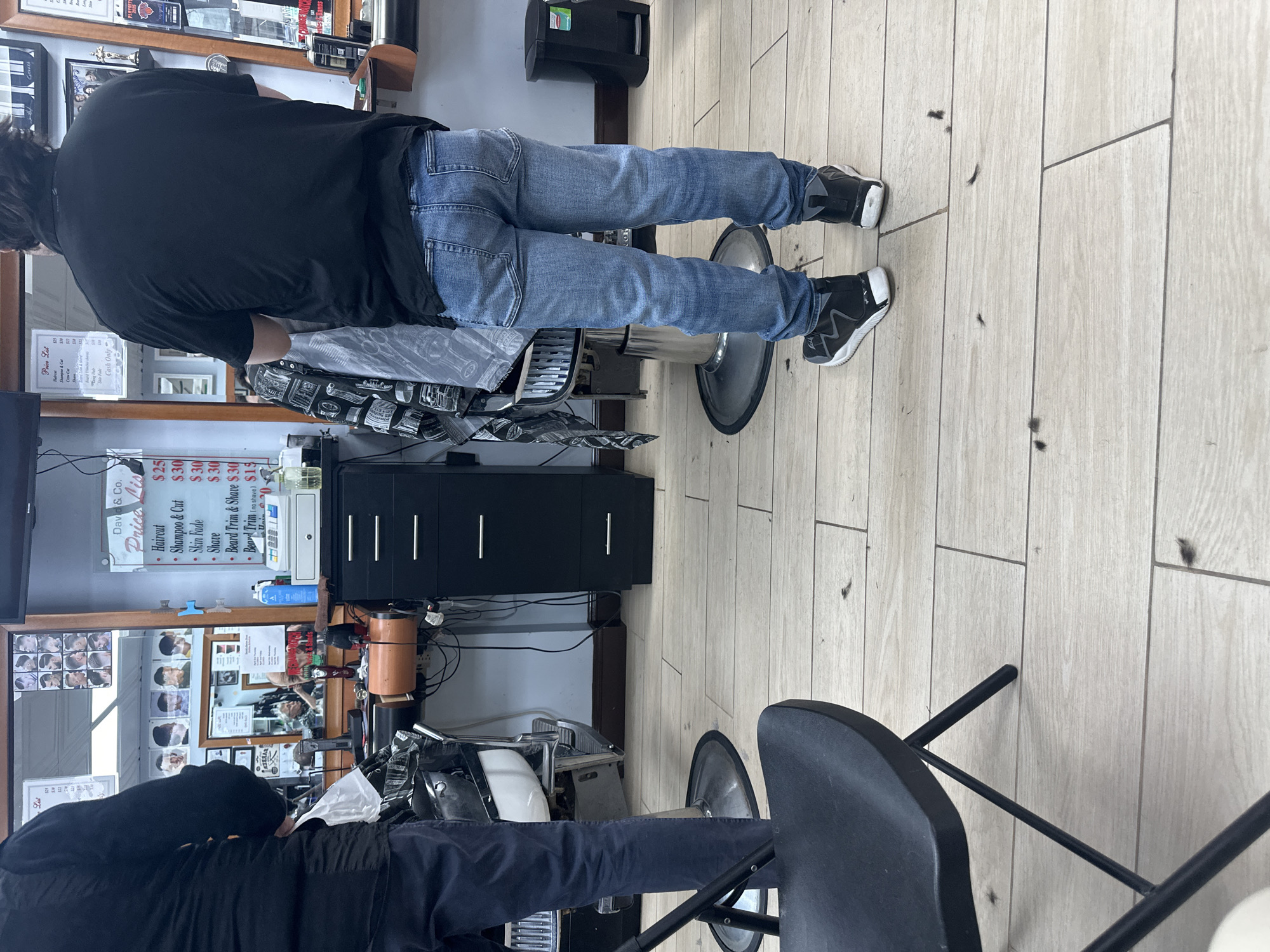 This image shows the interior of a barbershop. Two male figures are visible, one seated in a barber chair and the other obscured by a mirror, likely a barber. The person in the barber chair is wearing a black shirt and blue jeans, with black and white sneakers. Hair clippings are scattered on the light-colored, wood-look tiled floor.
Behind the barber chair is a black cabinet with three drawers. A price list for services, including haircuts, shampoo, skin fades, shaves, and beard trims, is visible on the wall. The price list indicates "David & Co." as the establishment's name. The list also states "Cash Only".
The image does not provide specific details to definitively place the barbershop within New York City, United States of America.