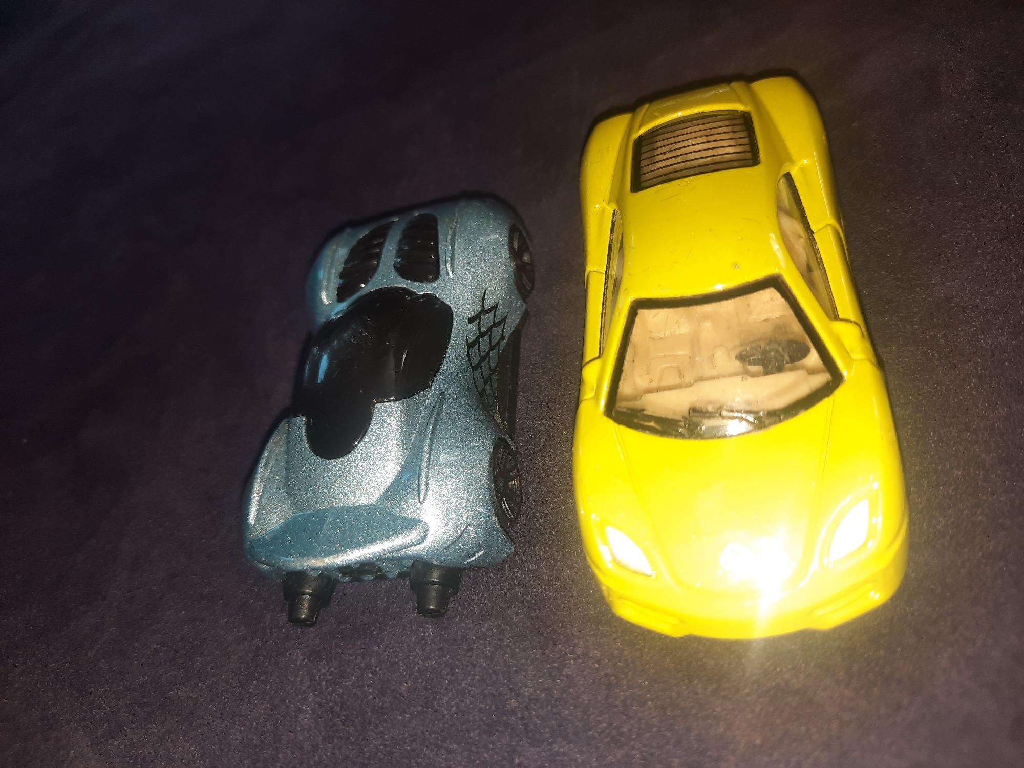 Two toy cars are arranged on a dark, textured surface. The upper car is a light blue, futuristic-looking vehicle with black accents and two visible exhaust pipes at the rear. It has a distinctive web-like pattern on its sides. The lower car is a bright yellow sports car with a transparent windshield and interior visible. Its headlights are illuminated, casting a bright glow. The scene is lit from above and slightly to the right, creating highlights and shadows on the objects and the surface. No people or specific locations are identifiable in this image.