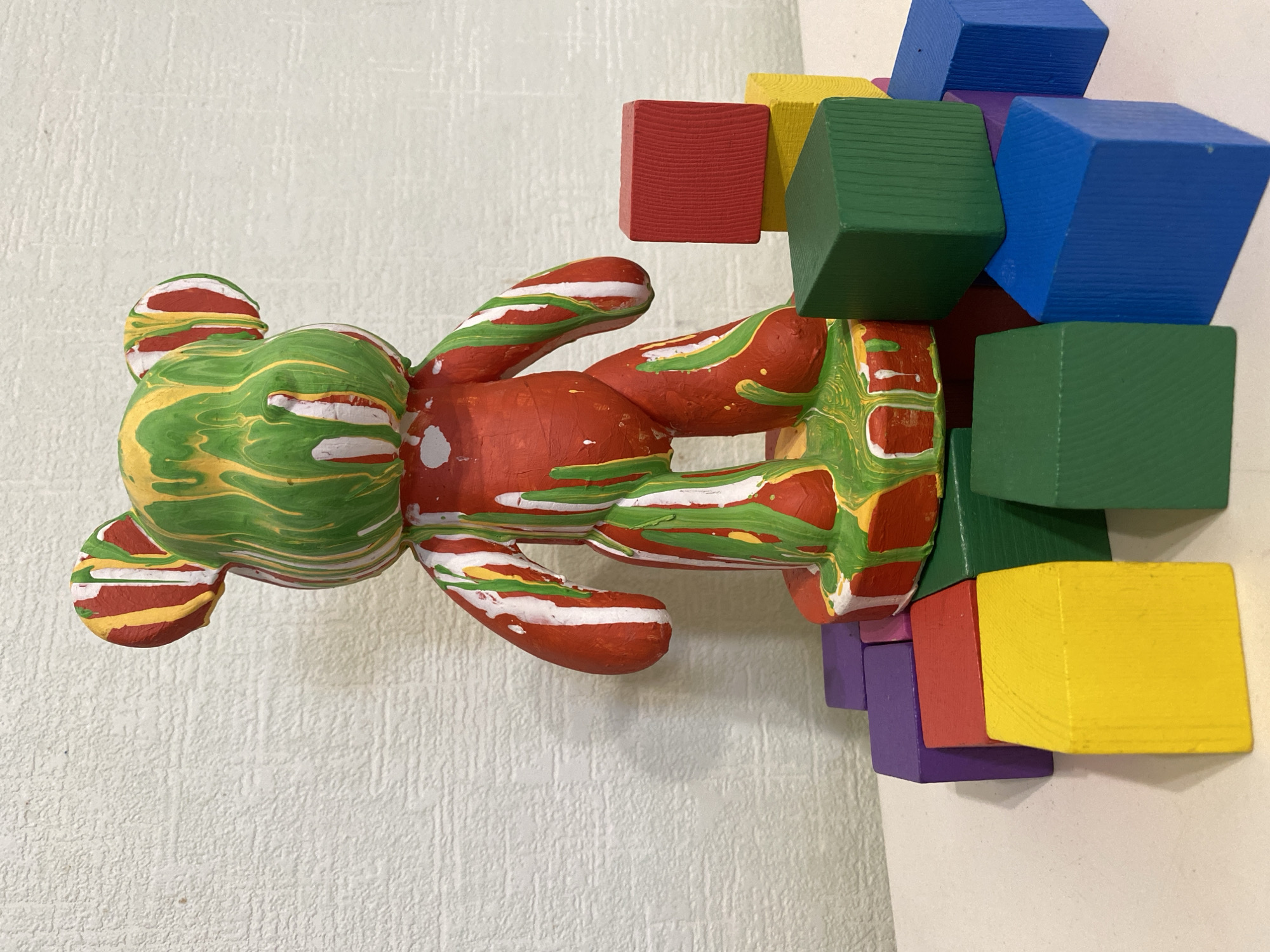 A decorative figurine is pictured against a textured off-white wall and an off-white background. The figurine is a bear-like shape, lying on its side with its head facing left. The bear has a marbled appearance, predominantly green and yellow on the head and red on the body and limbs, with white streaks throughout. Arranged around the right side of the bear's body are several small, painted wooden cubes in various colors, including blue, green, yellow, red, and purple. The blocks appear to be stacked haphazardly against the bear.
There is no definitive information about the location in the city of Sochi, Russia, or the people in this image.