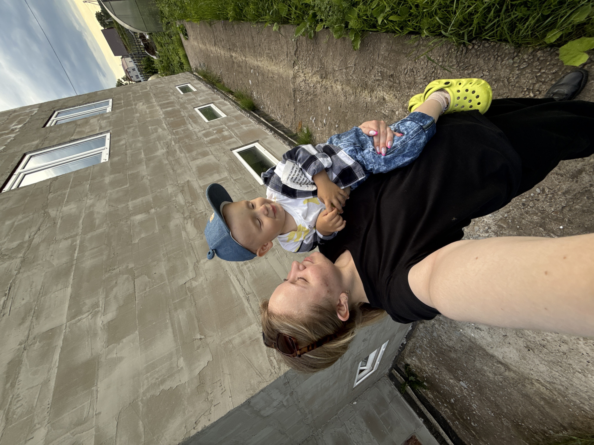 In this outdoor, low-angle selfie, a woman and a young child are the central subjects, positioned in what appears to be a construction or unfinished residential area. The woman, with blonde hair and sunglasses perched on her head, lies on her back, looking up at the child. The child, wearing a blue cap and a plaid shirt over a white graphic tee, is lying on the woman's chest, also looking down at her. The child's legs are bent, with one foot in a bright yellow Croc and the other bare.
The background features a partially constructed building with a concrete facade and several small, rectangular windows. Beyond the building, green grass and foliage are visible, with some houses and a greenhouse further in the distance. The sky above is partly cloudy, suggesting it's daytime, possibly late afternoon given the warm light. The concrete pathway and ground indicate an outdoor setting, likely in a rural or suburban area. The woman is wearing a black top and a long black skirt or dress, while the child is in blue jeans. The overall impression is a tender moment between a mother and child, captured casually amidst an ongoing construction project.
Visible text from a garment reads: "WHEN THE SUN COMES IF AND YOU WAKE UP KNOW THAT YOUR BE FULL OF AD".