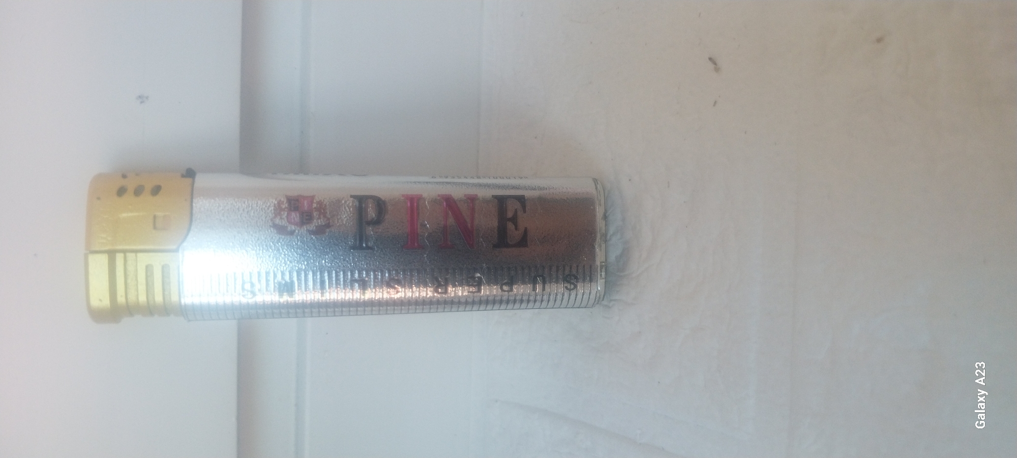 A silver lighter with a gold top stands upright on a white surface. The lighter has a textured metallic finish and features red text that reads "PINE" vertically down one side. There are also subtle markings that appear to be measurement lines along the side of the lighter. The background is a white, horizontally-lined surface, likely a wall or partition. The bottom left corner of the image shows "Galaxy A23" indicating the device used for capture. The scene is clean and well-lit, with no visible signs of damage or distress. There are no people present. The specific location within Tuapse, Russia, cannot be determined from the provided image.
