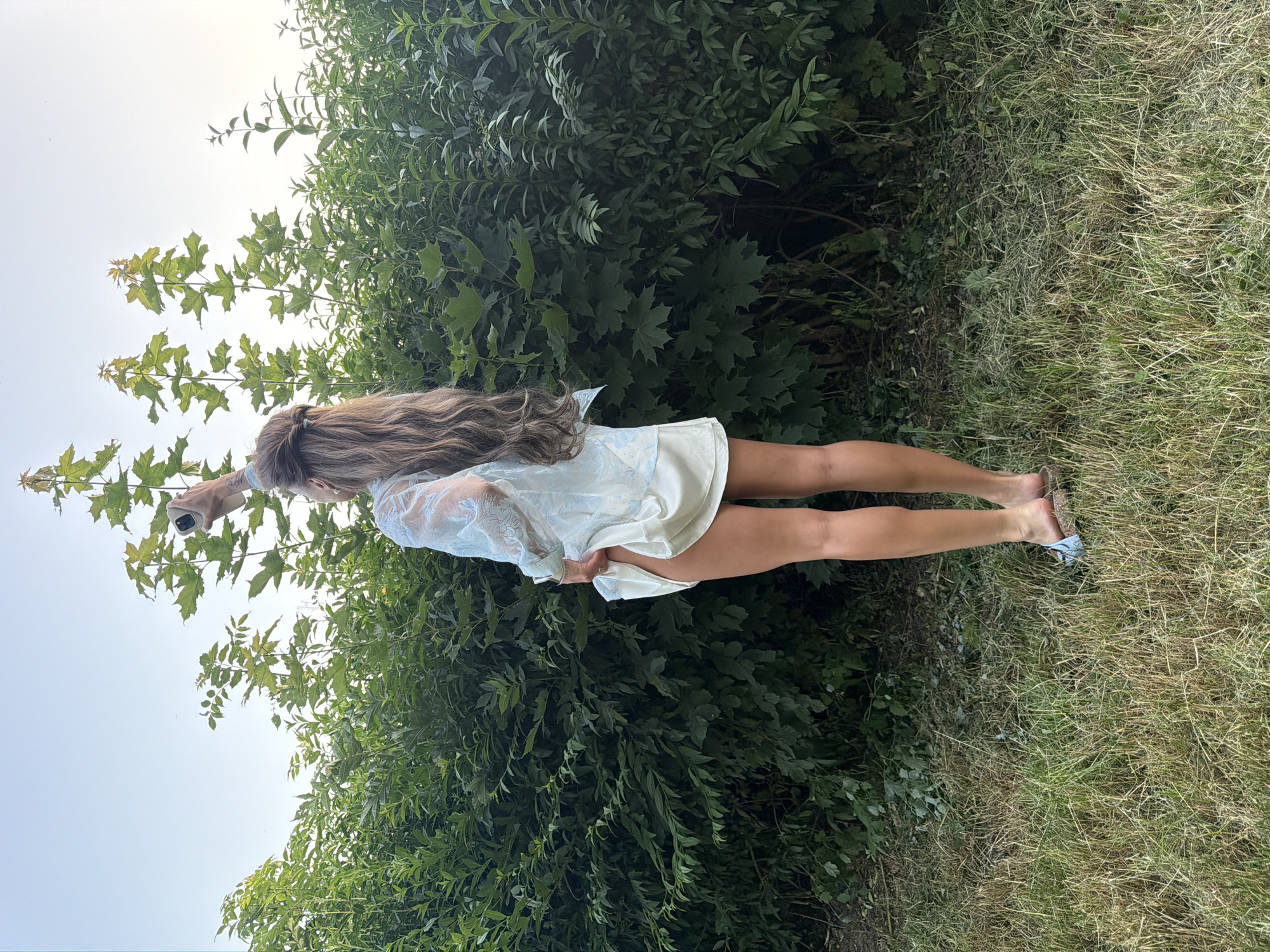 The image shows a woman standing in front of a dense green hedge, looking up and holding a phone in her right hand. She is wearing a light blue sheer top with lace details and a white slip dress underneath. Her long brown hair is styled in a messy bun with some strands falling loose. The hedge forms a natural backdrop, with tall leafy trees extending upwards into a clear sky. The ground in front of the hedge is covered in dry grass. The woman's legs are visible, wearing light blue sandals. The scene appears to be outdoors, possibly in a park or garden. There is no indication of the specific location within Bila Tserkva, Ukraine.