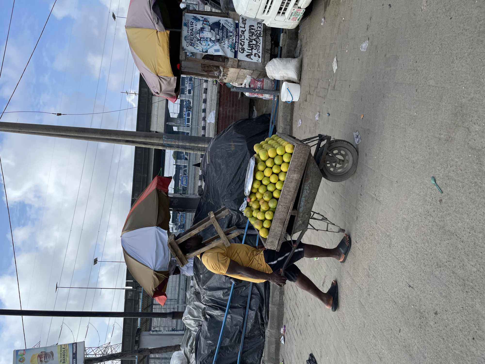 In Lekki, Nigeria, a man in a yellow t-shirt and black shorts pushes a wheelbarrow filled with oranges. He is walking on a paved surface, with a wooden stool balanced on his head. Behind him, there are several stalls with umbrellas, black tarpaulin covering goods, and signs. One sign advertises "CELL PHONE REPAIR SERVICE" and "Aluminum Glass." A white van with a license plate is parked to the right. The environment appears to be an outdoor market or street scene, with a cloudy sky and overhead power lines visible. Debris is scattered on the ground.