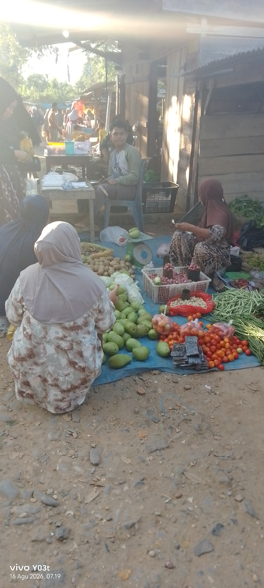 The image depicts an outdoor market scene, likely in Palosilae, Indonesia, during daylight. The ground is covered with dirt and scattered stones. In the foreground, a person wearing a patterned dress and a headscarf is seated on the ground, facing away from the camera, with their hands near a pile of green mangoes. To the left, another person in dark clothing and a headscarf is also seated. Various vegetables are spread out on a blue tarp: piles of potatoes, cabbages, green mangoes, tomatoes, and long green beans. A weighing scale is visible with green mangoes on it. A white basket contains red onions and another contains small white nuts. Behind the seated figures, a young person in a light-colored shirt with blue overalls sits on a plastic chair, looking towards the left. Further in the background, stalls with wooden structures and corrugated metal roofs are visible, along with other people browsing or working at the market. The overall atmosphere suggests a typical local market environment.