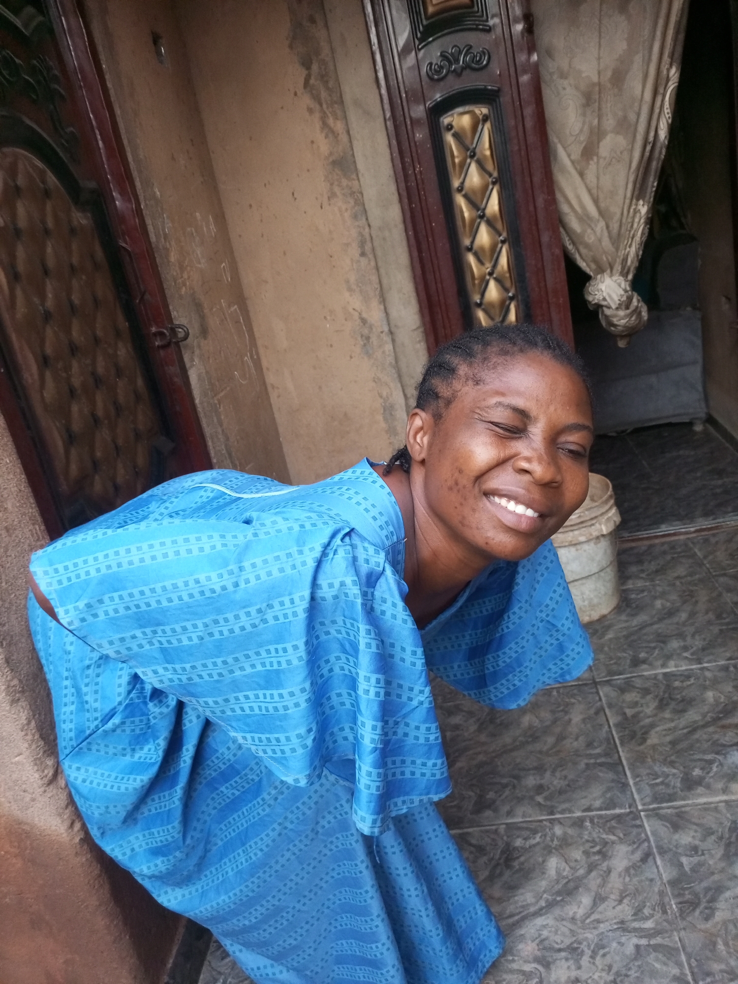 An eye-level, medium shot captures a woman with dark skin, braided hair, and dark eyes in Tempure, Nigeria. She is wearing a blue dress with a repeating square pattern. Her eyes are closed, and she is smiling widely, with her head tilted back slightly. She is leaning forward, with her left shoulder prominently visible. Behind her, a dark brown door with gold detailing is partially visible on the left. To the right, a beige curtain hangs, and a white bucket is situated on the tiled floor. The woman's posture and expression suggest a moment of joy or laughter.