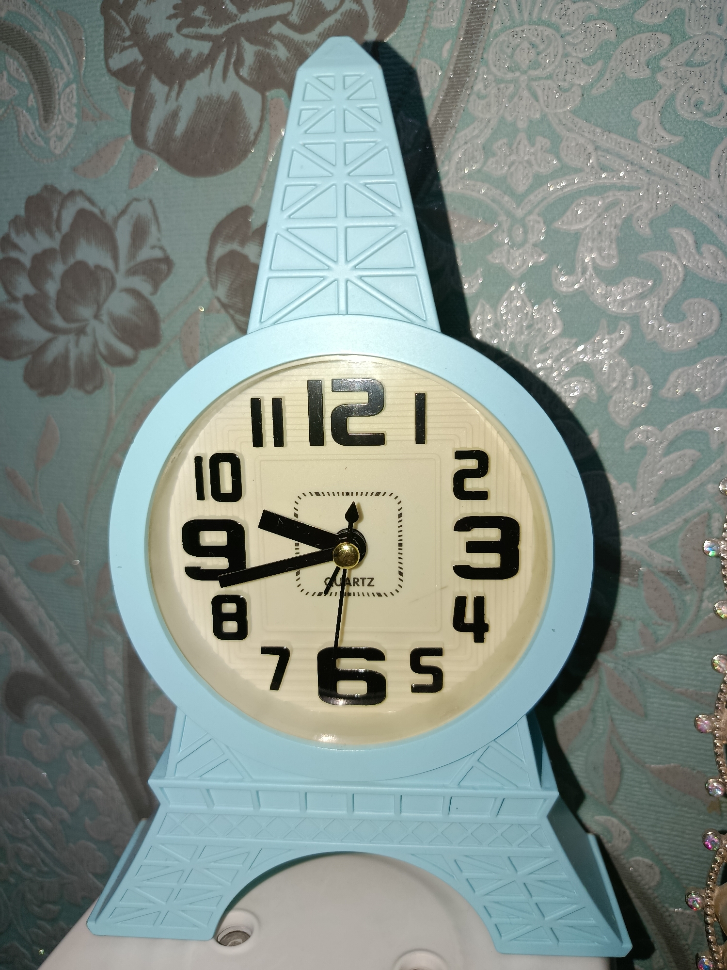 The image shows a light blue alarm clock in the shape of the Eiffel Tower. The clock face is round with black numbers and hands. The hour hand is pointing between 9 and 10, and the minute hand is pointing at the 5. The background is a textured wallpaper with a floral pattern in shades of blue and grey. The clock is placed on a white surface. The scene is indoors. There are no people or animals visible in the image. There is no information in the image to suggest it is located in Tashkent, Uzbekistan.