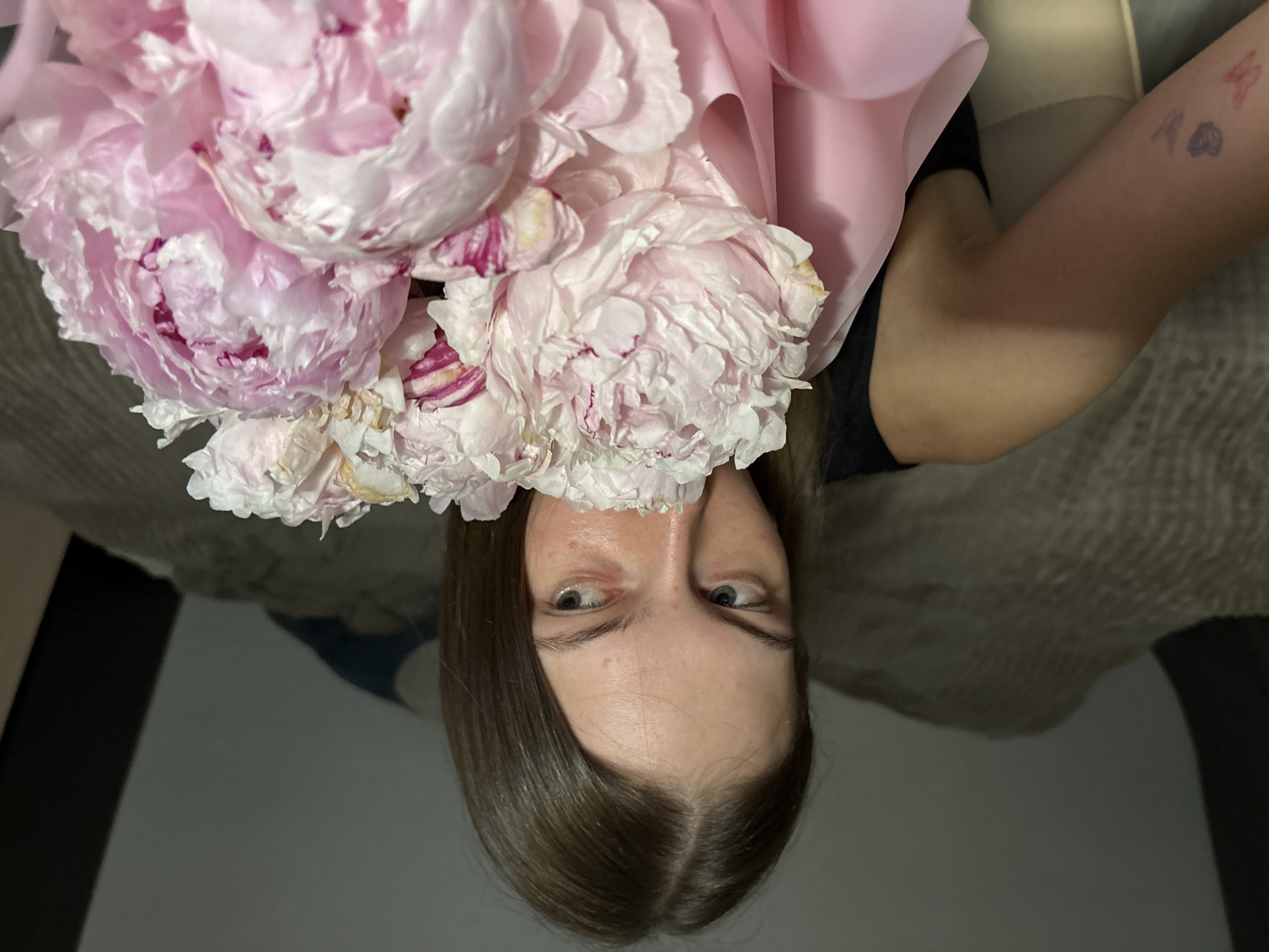 A young woman with long brown hair is holding a bouquet of pink peonies. She is looking to her right, with her face partially obscured by the flowers. Her left arm is visible, and there are temporary tattoos of a heart, a butterfly, and what appears to be the number "2" on her forearm. She is wearing a black tank top. The background is blurred, but appears to be an indoor setting, possibly a bedroom, with a beige textured surface that could be a blanket or duvet. The lighting is dim. The image was taken in Perm, Russia.