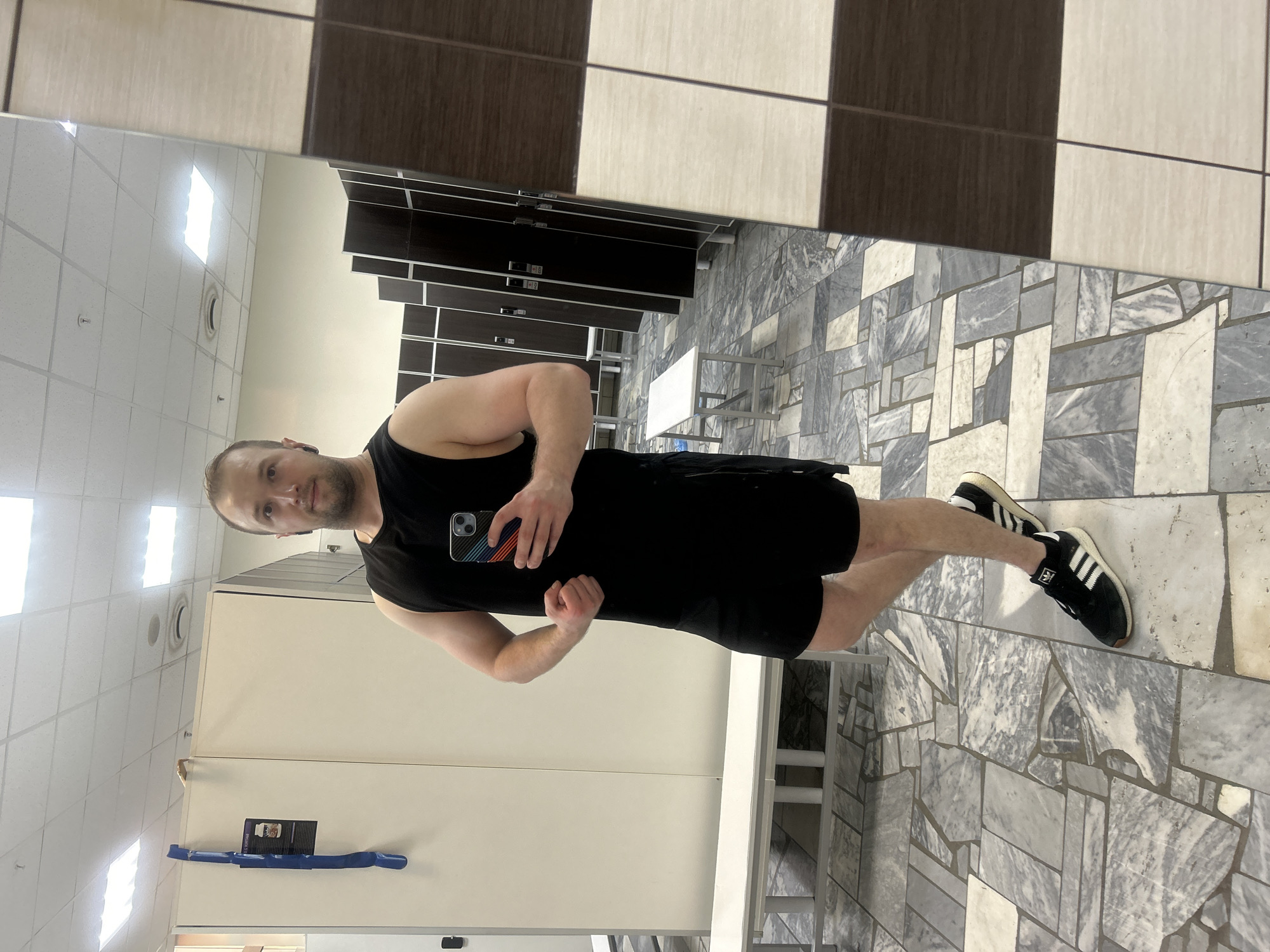 A man in a black tank top and shorts is posing in a mirrored locker room with black and white tile, presumably in Chaikovsky, Russia. He holds a cell phone with a multi-colored case in his left hand, while flexing his right arm. He is wearing black Adidas sneakers with white stripes. Lockers are visible behind him, and a marble-patterned floor is present. A white table is behind him, attached to the wall. A blue exercise band is mounted on the wall to the left.