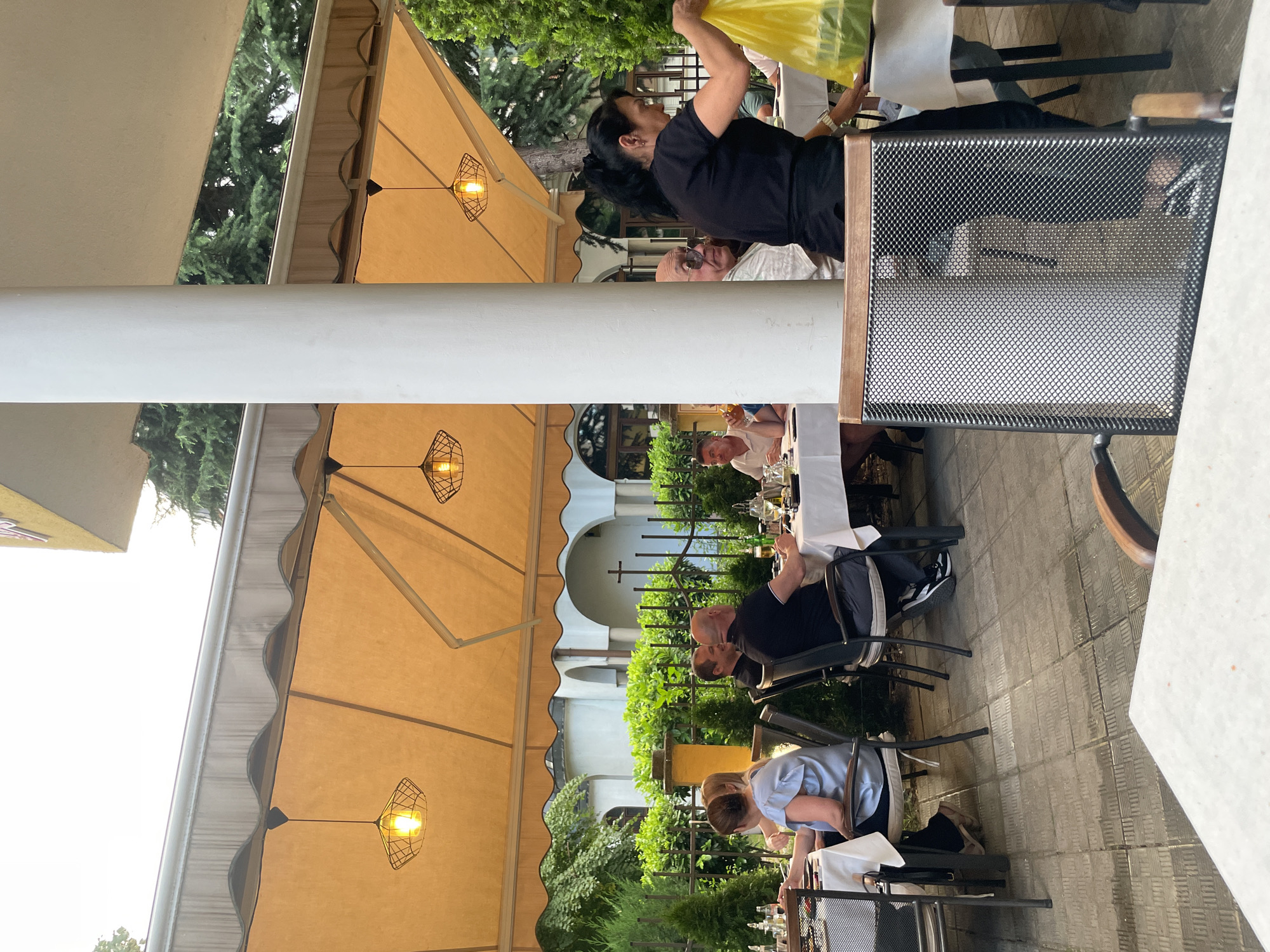 An outdoor restaurant patio scene features a group of people dining at tables under a large, orange awning. In the foreground, a woman in a black shirt is holding a bright yellow plastic bag. Behind her, patrons are seated at white-clothed tables, some engaged in conversation. The tables are set with plates, glasses, and bottles. In the background, a fence with lush green foliage separates the patio from a building with arches and a cross, suggesting a church or similar structure. The ground is paved with square tiles. Decorative light fixtures hang from the awning.