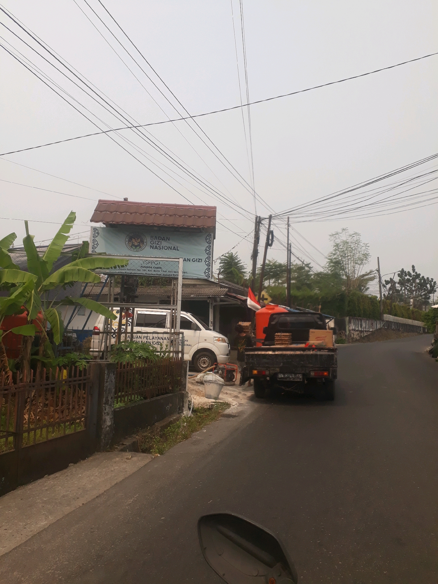 The image depicts a street scene in Pangkalpinang, Indonesia, characterized by an overcast sky and a road lined with buildings and vegetation. In the foreground, a motorcycle's rearview mirror is visible. The road itself is paved and appears somewhat wet. To the left, a dark metal fence borders a property with a concrete sidewalk. Behind the fence, banana trees and other foliage are present. A white van is parked in front of a building with a blue banner that reads "BADAN GIZI NASIONAL" (National Nutrition Agency) and "PONDOK CABIK". A black pickup truck with an orange water tank and some boxes is parked on the right side of the road, facing away from the camera. A person is standing near the rear of the pickup truck. The environment suggests a slightly hazy or foggy day, with numerous overhead power lines crisscrossing the sky.