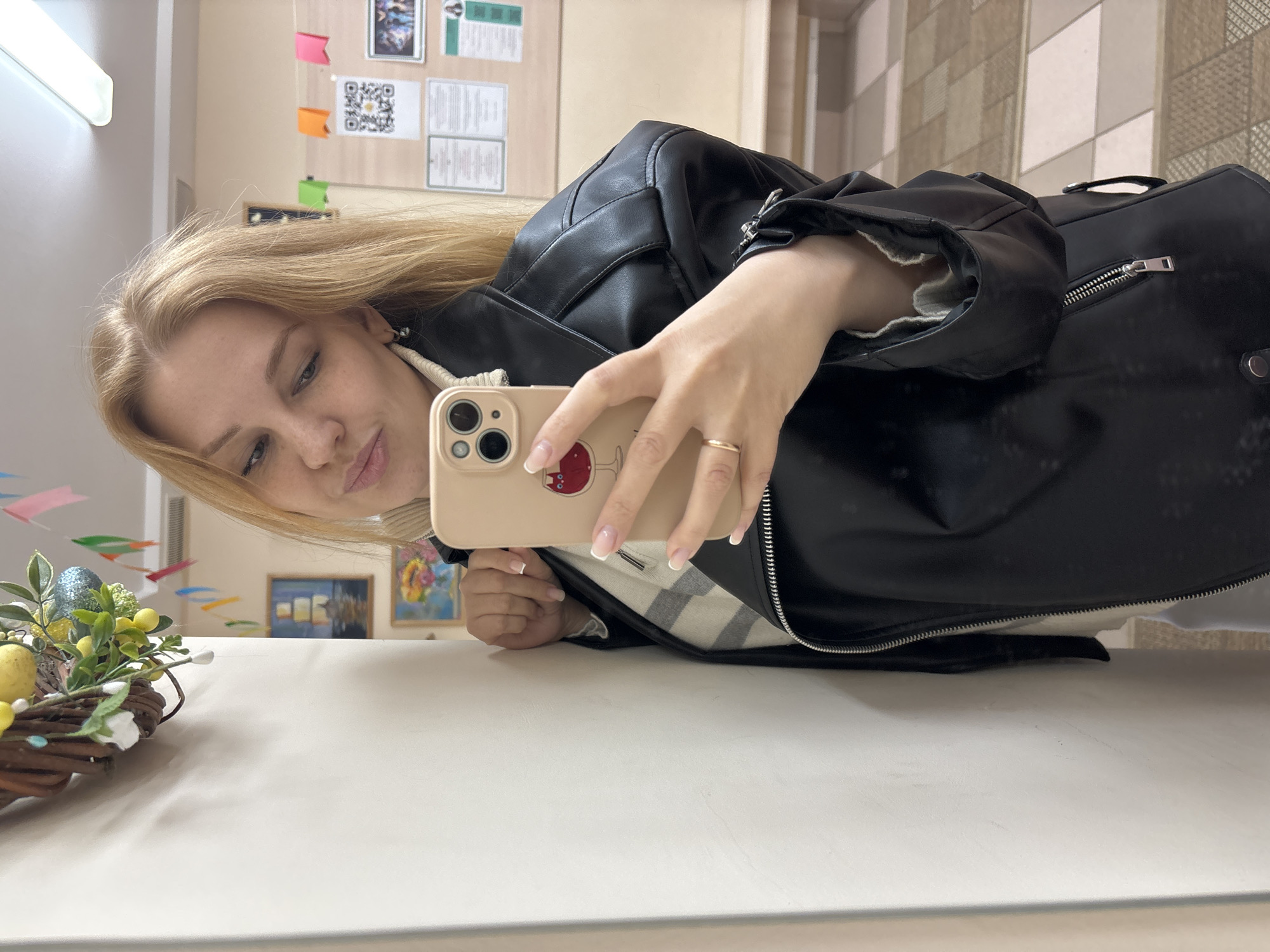A young woman with blonde hair and fair skin is taking a selfie in a room that appears to be reflected in a shiny surface, likely a table. She is wearing a black leather jacket and a sweater with a white and grey stripe pattern. She is holding a beige-colored phone with a case decorated with a cartoon. The background includes a bulletin board with pinned papers and colorful sticky notes, suggesting an office or school environment. In the reflection on the table, there is an Easter wreath with eggs and flowers, indicating a festive or decorative touch. There are no explicit visual indicators to confirm the location as Podosenikha, Russia.