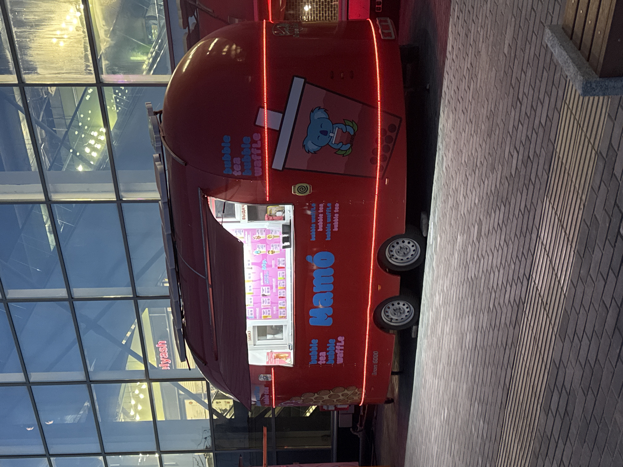 A bright red food truck, branded "Mamó," is parked on a brick-paved walkway in front of a modern building with large glass windows. The food truck has a rounded design and is decorated with red LED lights along its base and around the top edge of its canopy. On the side of the truck, the words "bubble tea," "bubble waffle" are displayed, along with a graphic of a koala holding a cup. A menu is visible inside the service window, featuring items like "bubble tea" and prices in Russian currency. The building's reflective windows show an interior with bright lights and escalators, suggesting it's a commercial or public space. The overall scene depicts an outdoor food vendor setting up in an urban environment, possibly during the evening or at dusk, as indicated by the artificial lighting. The location is identified as Novosibirsk, Russia.