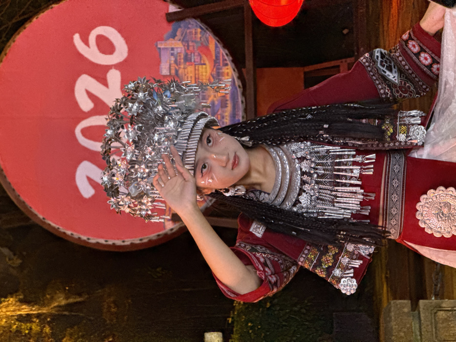 This image, captured in Yuzhong District, China, features a young individual, likely female, dressed in elaborate traditional attire during nighttime. The subject stands centrally, looking directly forward, with a solemn expression. She wears a prominent silver headdress composed of layered bands, radiating star-like motifs, and numerous small dangling ornaments. Her hair is styled in multiple thin braids cascading over her shoulders. Her face is made up with white foundation, highlighted by small red markings below her eyes and on her upper cheekbones. Adorning her neck is a multi-layered silver necklace with a wide collar and intricate dangling pendants, complemented by matching earrings. Her attire consists of a dark red tunic with broad sleeves, detailed with embroidered patterns in red, black, and white on the cuffs and front panels. A white undergarment is visible beneath the tunic and as a skirt. A large, circular silver medallion with embossed patterns is affixed to the front of her red skirt. Her right hand is raised, palm facing outward, with fingers slightly spread, positioned near her temple. In the background, a large, circular red backdrop is visible, featuring the white numbers "2026" prominently displayed. Below these numbers, a colorful illustration of a multi-tiered traditional cityscape is depicted on the lower right portion of the backdrop. The scene is illuminated by artificial light, casting reflections on the silver ornaments. Dark, indistinct foliage or architectural elements are present in the left background, and a blurred red light source is partially visible on the right.