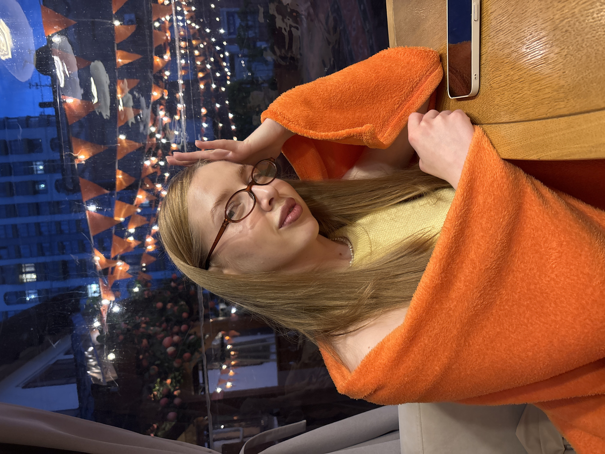 A young adult female is depicted within an interior setting consistent with a restaurant or cafe in Perm, Russia, during twilight conditions. She has long, straight, blonde hair parted on the side and wears oval-shaped, brown-rimmed glasses. Her left hand is raised, with fingers splayed, resting against her forehead. She is wearing a light yellow, possibly ribbed, sweater or top and a pearl necklace. Her upper body is draped in a bright orange, fuzzy, fleece-like blanket or shawl that covers her shoulders and arms. Her right forearm and hand rest on a light-colored wooden table. A white smartphone with a visible camera array lies face down on the table to her right. Through a large window behind her, an urban exterior is visible under a deep blue twilight sky. Distant dark buildings display illuminated windows. The exterior features multiple strings of white and yellow lights and a horizontal arrangement of orange triangular pennants. Below these decorations, a structure covered with numerous red, spherical objects resembling apples is present.