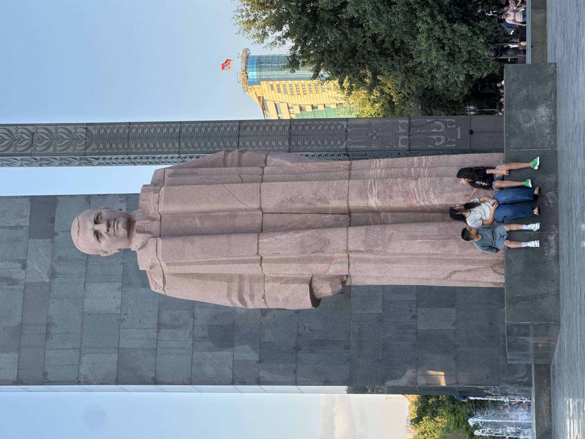 The image captures a view of the Mother Armenia statue in Yerevan, Armenia. In the foreground, three children are seated on a stone platform in front of the statue's massive base. A young boy in a grey t-shirt and shorts sits on the left, while two girls in t-shirts and jeans occupy the center and right positions. Behind them, the statue itself looms, depicting a woman in a robe. To the right of the statue, a tall building with a prominent flag flying from its top is visible amongst the green trees. The scene is bathed in the soft light of what appears to be late afternoon or early evening. A fountain is visible to the left of the statue, with water spraying upwards. The ground in front of the statue is paved.